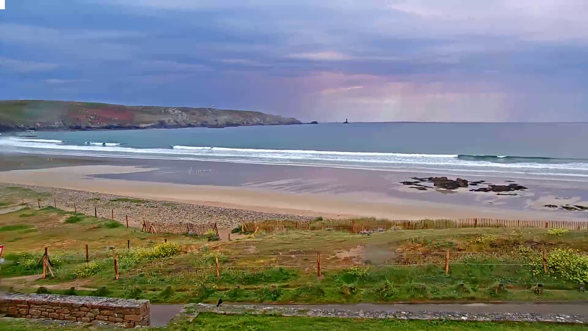 A sandy beach with gentle waves under a cloudy sky stretches to a distant headland and calm ocean.