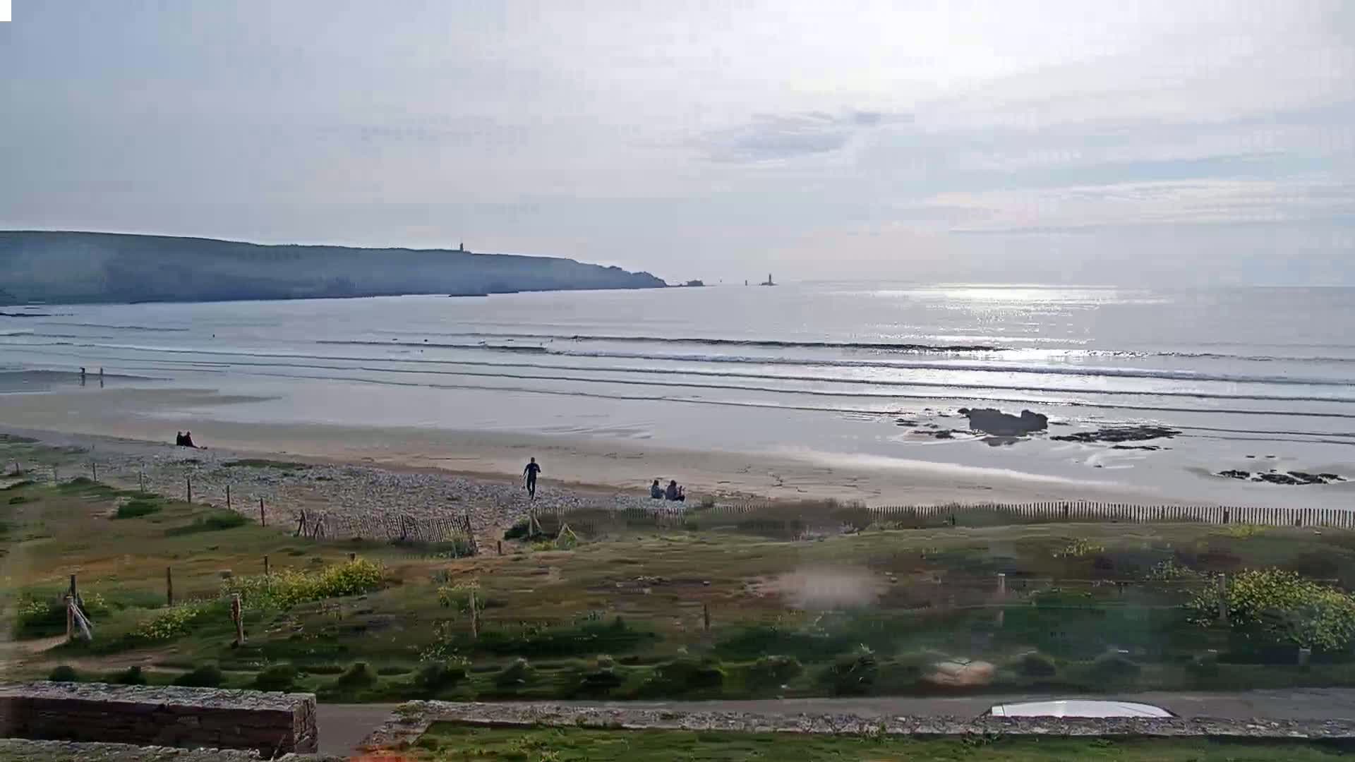 A sandy beach with gentle waves under a mostly clear sky, showing a few people and a distant coastline.