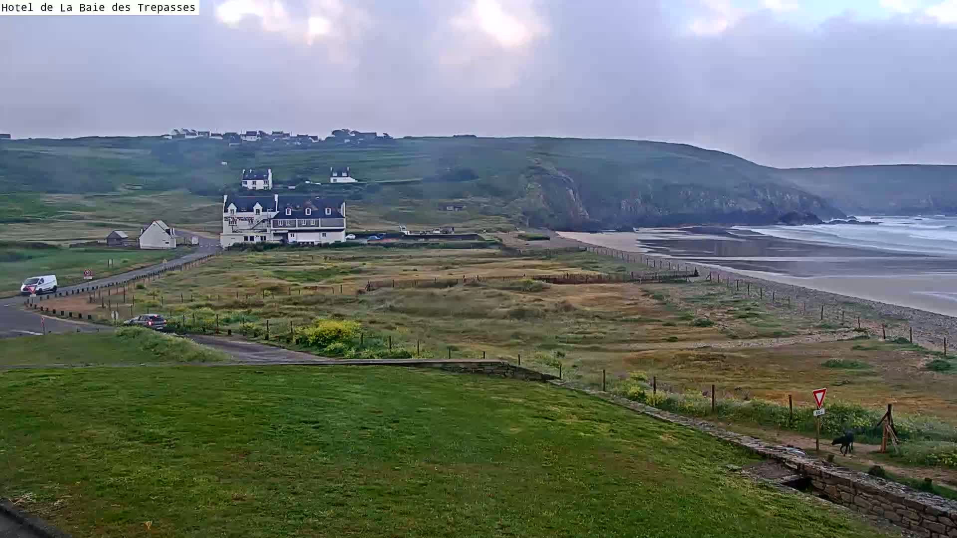 A coastal scene under an overcast sky shows a sandy beach with waves, a building, and a few cars near a grassy area leading to a road.