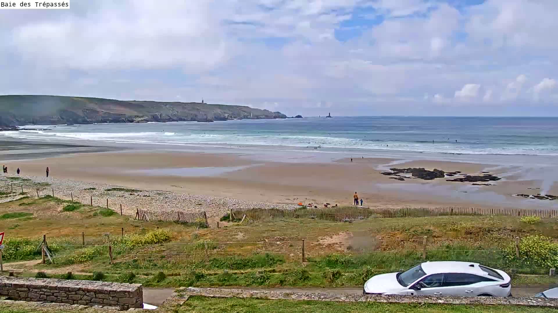 A sandy beach with gentle waves under a partly cloudy sky, a few people are visible on the beach and in the water, and a white car is parked nearby.