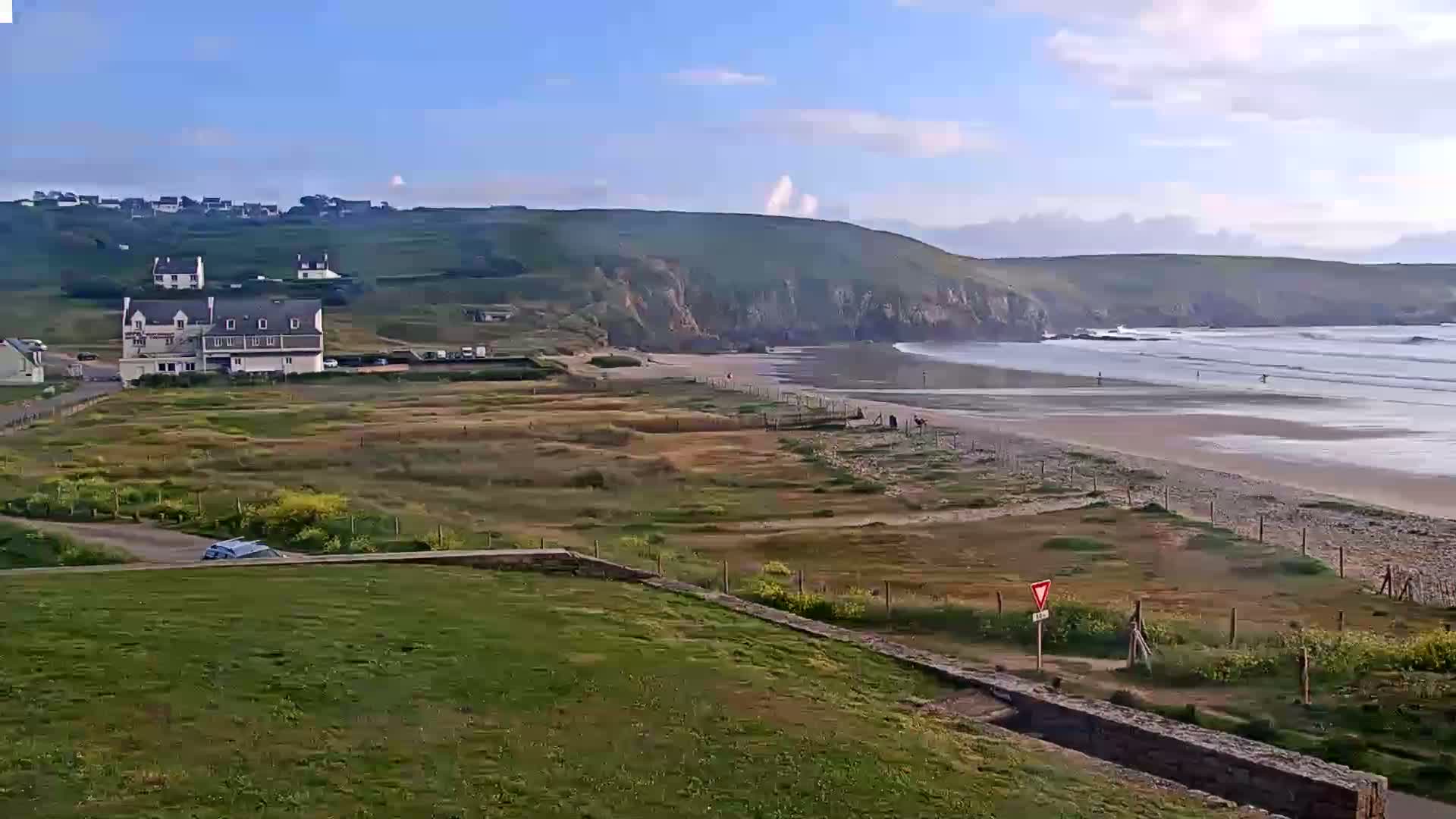 A sandy beach with gentle waves and a few surfers is visible below a grassy hill, where a small village and hotel are situated under a mostly clear sky.