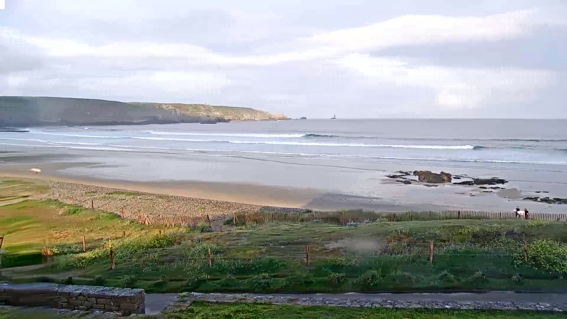 A wide sandy beach with small waves under a cloudy sky is viewed from a grassy hilltop.