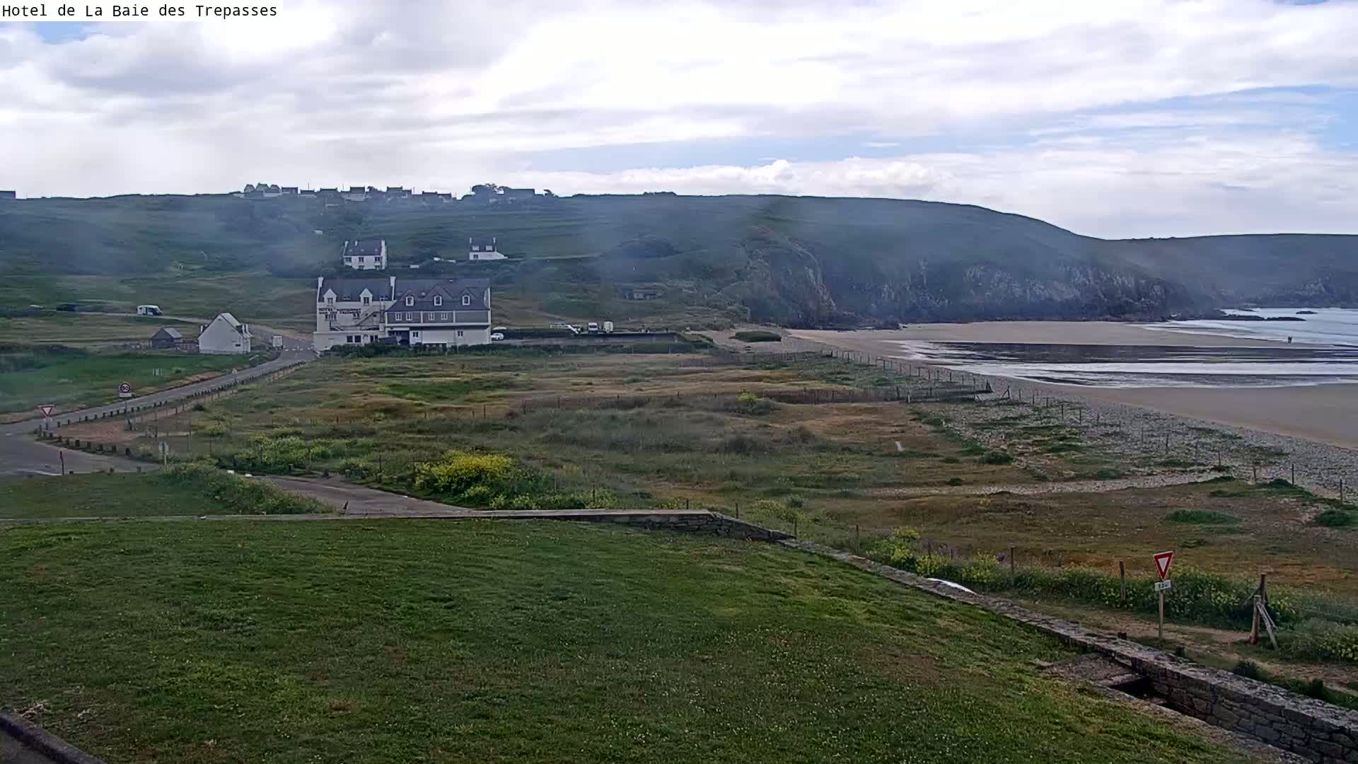 A coastal scene shows a sandy beach, a grassy area, and a cluster of buildings near a cliff under a cloudy sky.