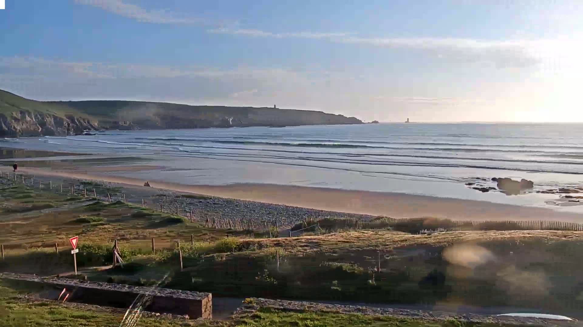 A sandy beach with gentle waves rolls onto the shore under a clear, bright sky, backed by grassy hills and a distant coastline.