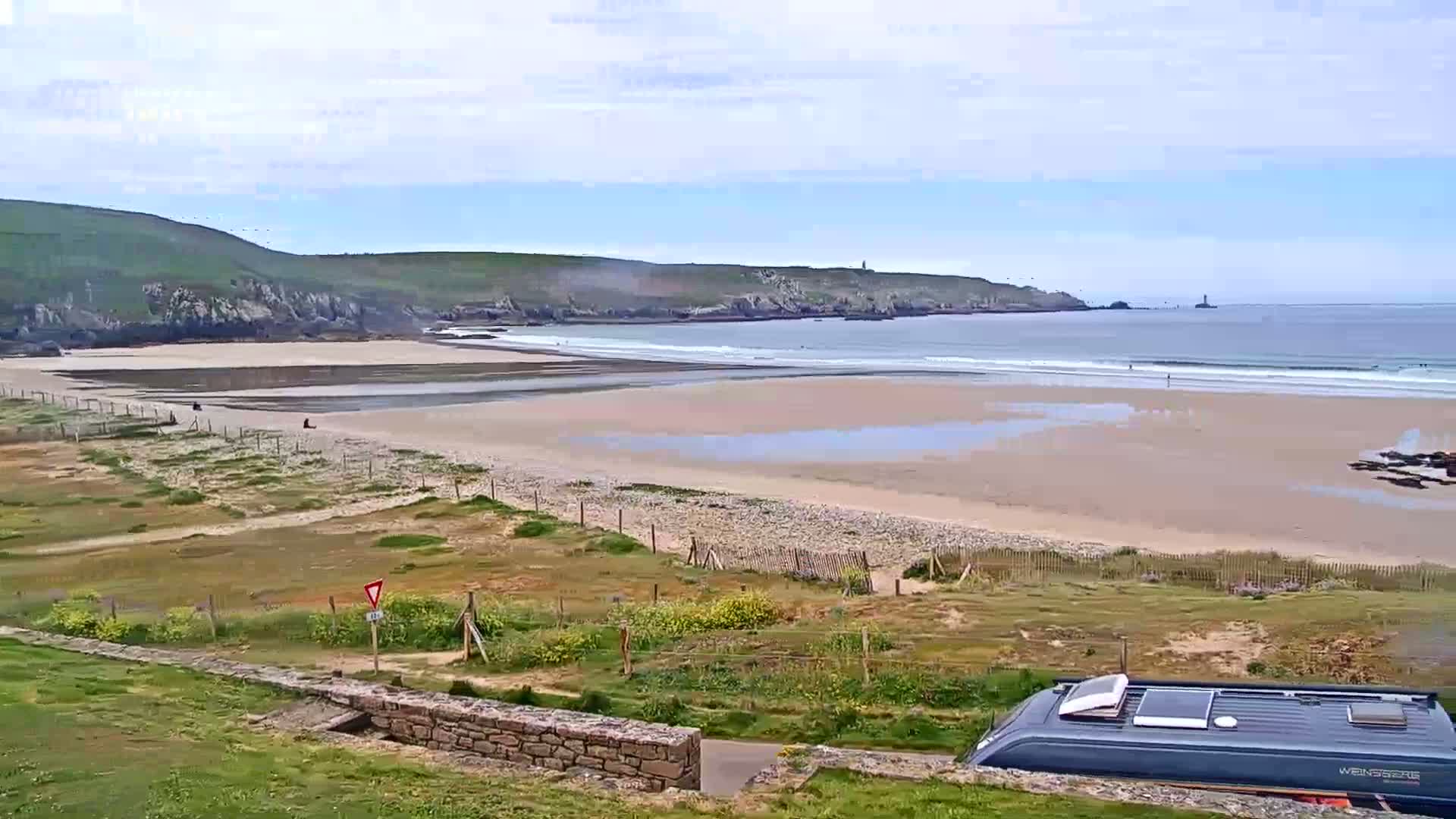 A sandy beach with small waves under a partly cloudy sky is viewed from a grassy hill overlooking a campervan.