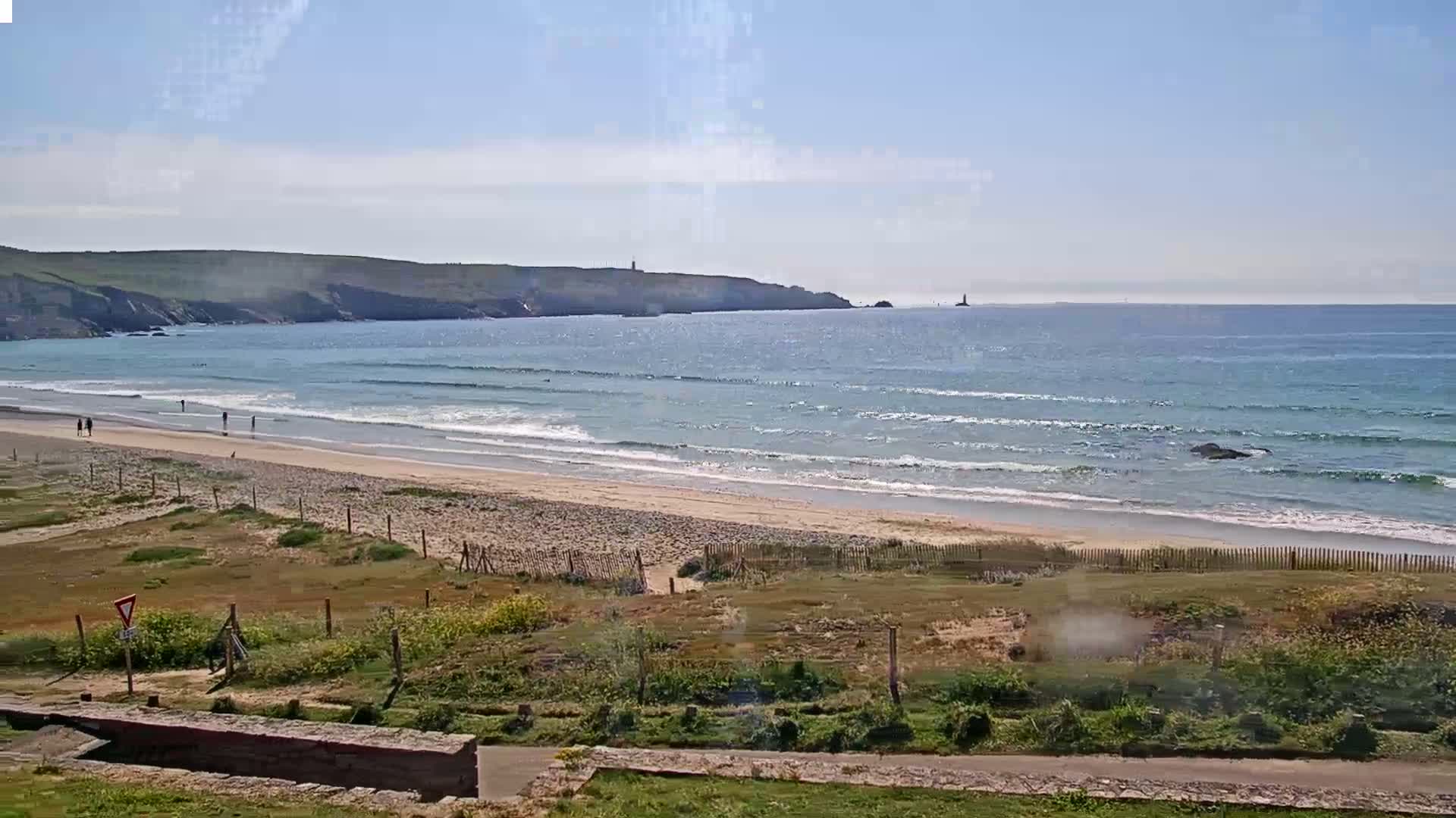 A sandy beach with gentle waves under a mostly clear sky, backed by grassy dunes and a distant rocky coastline.