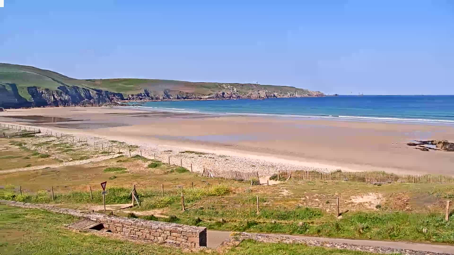 A sandy beach stretches out to a calm, blue ocean under a clear blue sky, backed by green hills.