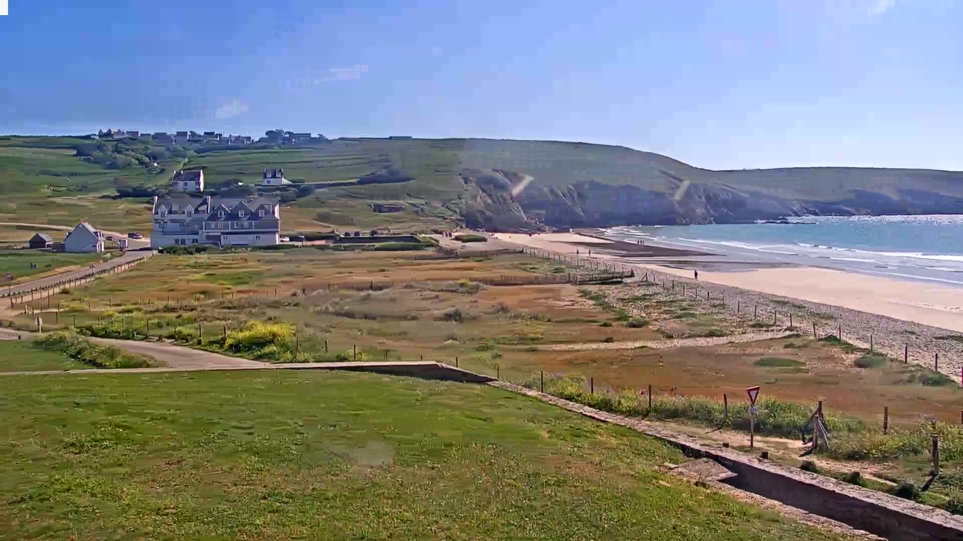 A sandy beach with gentle waves under a clear blue sky is visible from a grassy overlook, featuring a large building and a small village on a nearby hill.