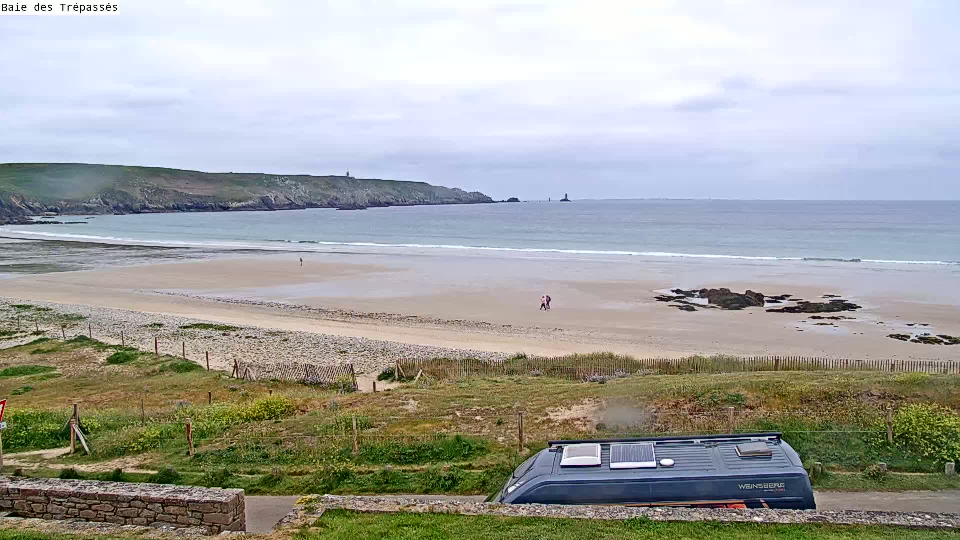 A sandy beach with gentle waves stretches below a cloudy sky, a camper van parked in the foreground, and a few people walking on the shore.