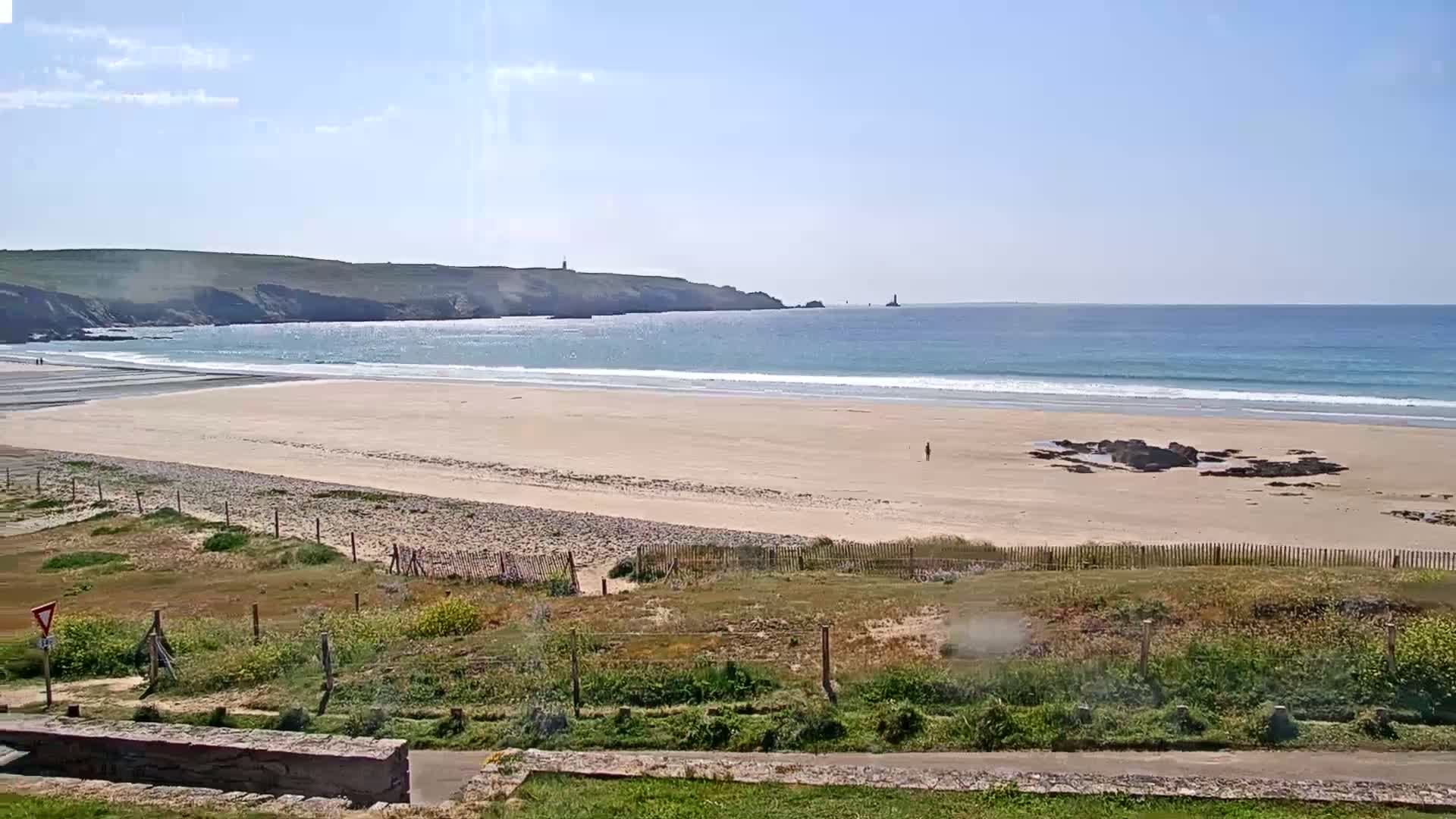 A sandy beach stretches out to a calm ocean under a bright, sunny sky, with green hills visible in the distance.