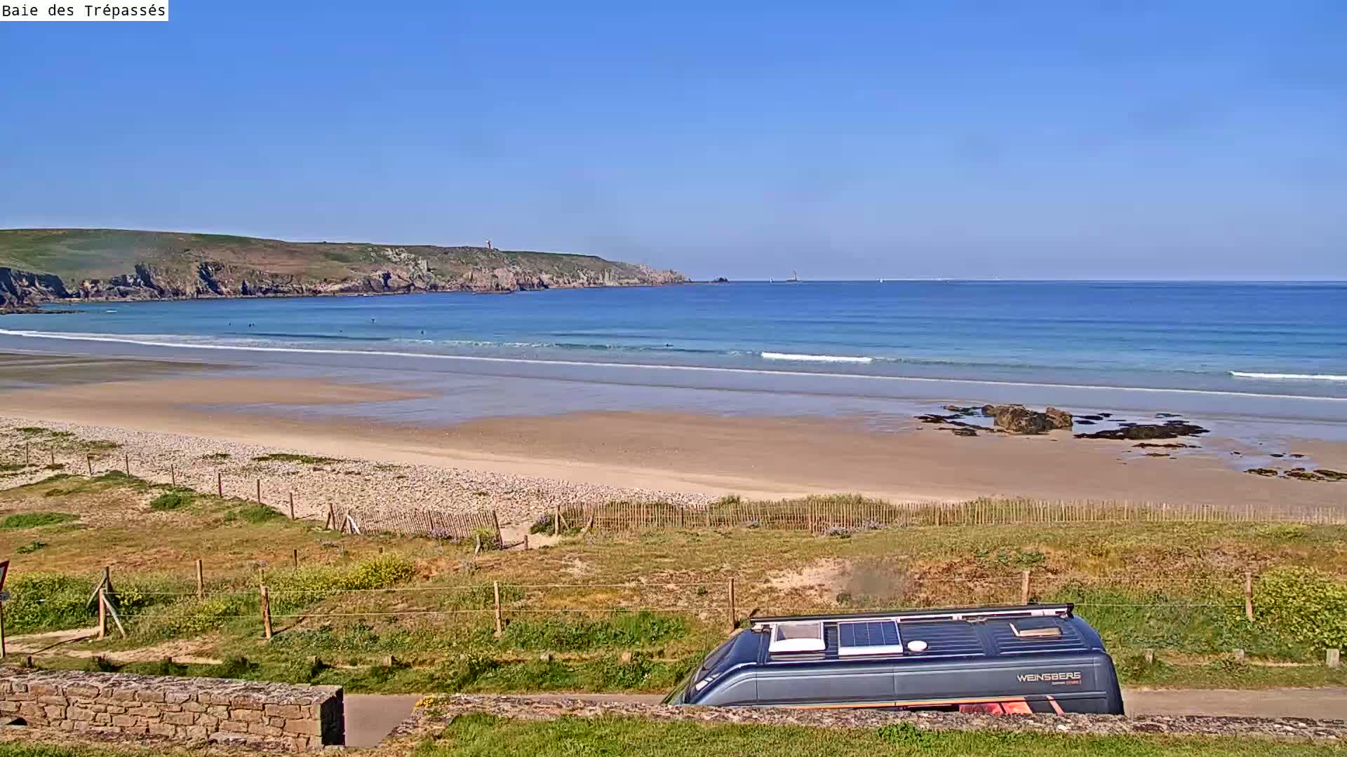 A camper van is parked overlooking a sandy beach with small waves lapping the shore under a clear blue sky.