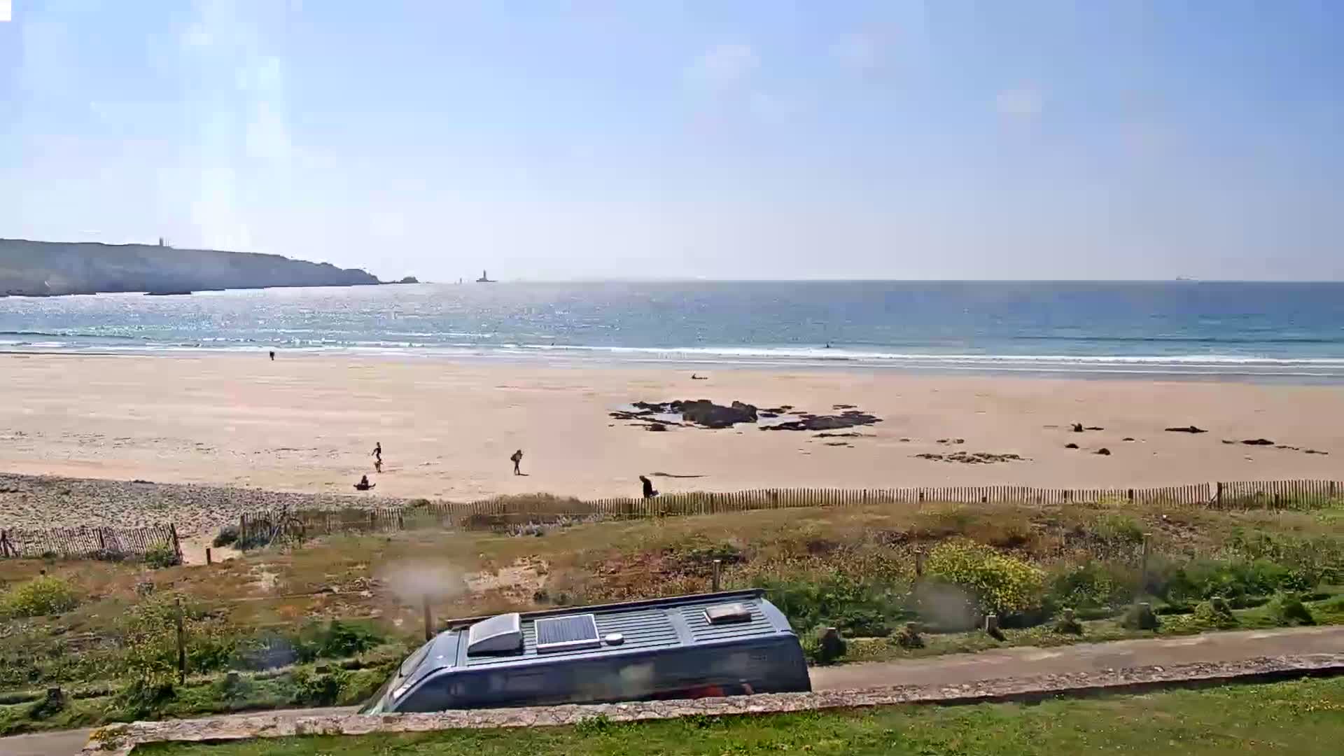 A sandy beach with a few people and a camper van is visible, overlooking a calm ocean under a sunny sky.