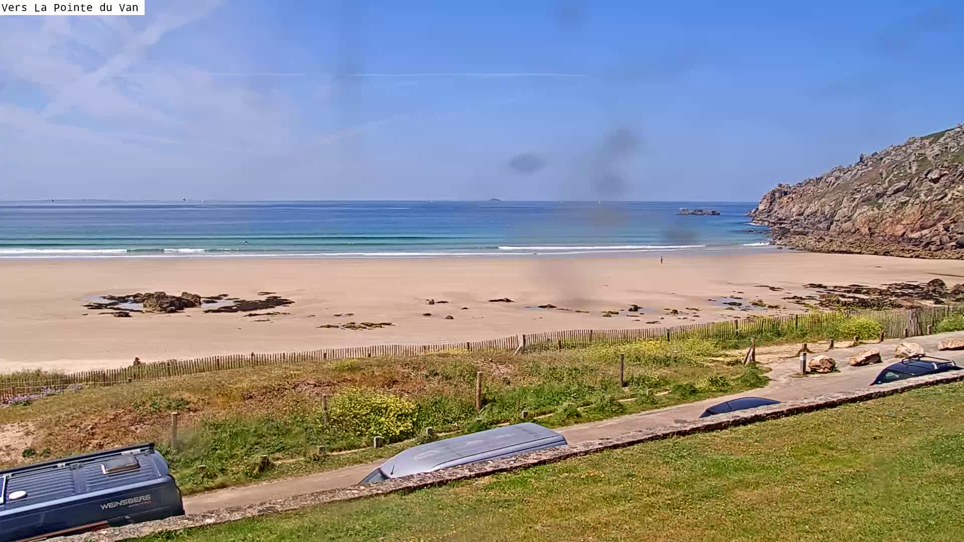 A sandy beach with small waves under a clear blue sky is seen from an elevated vantage point, with several vehicles parked along a road overlooking the scene.