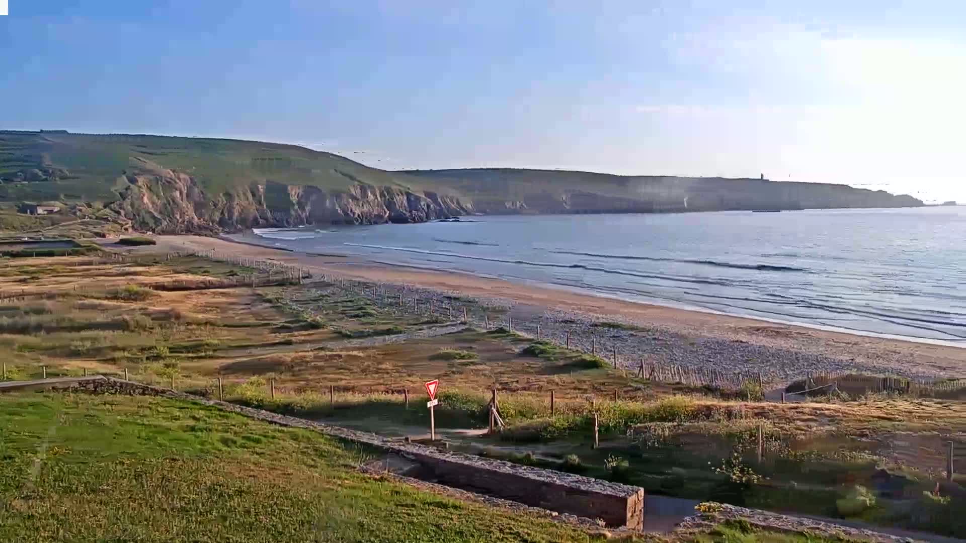 A sandy beach with gentle waves laps against the shore under a clear blue sky, backed by green hills and a calm ocean.