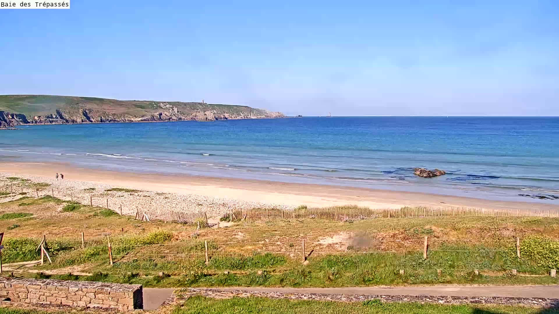 A sandy beach with gentle waves stretches out to a calm ocean under a clear blue sky, backed by a grassy headland.