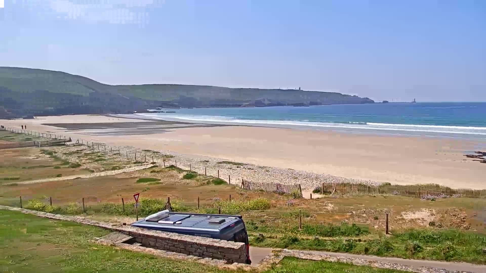 A sandy beach with gentle waves stretches beneath a clear blue sky, a van parked nearby, and a grassy area separating the viewer from the coast.