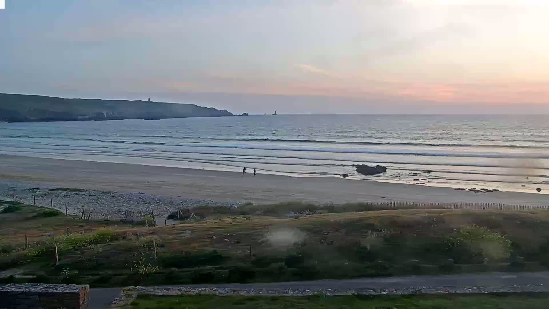 A sandy beach with gentle waves under a soft, pinkish sunset sky, with two people walking on the shore.