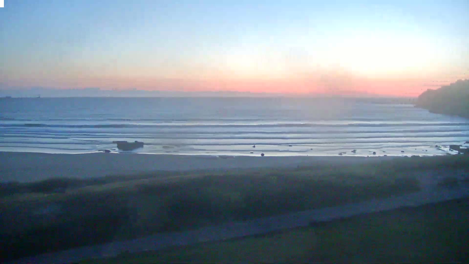 A sandy beach with gentle waves rolls onto the shore at sunset under a hazy, pink sky.