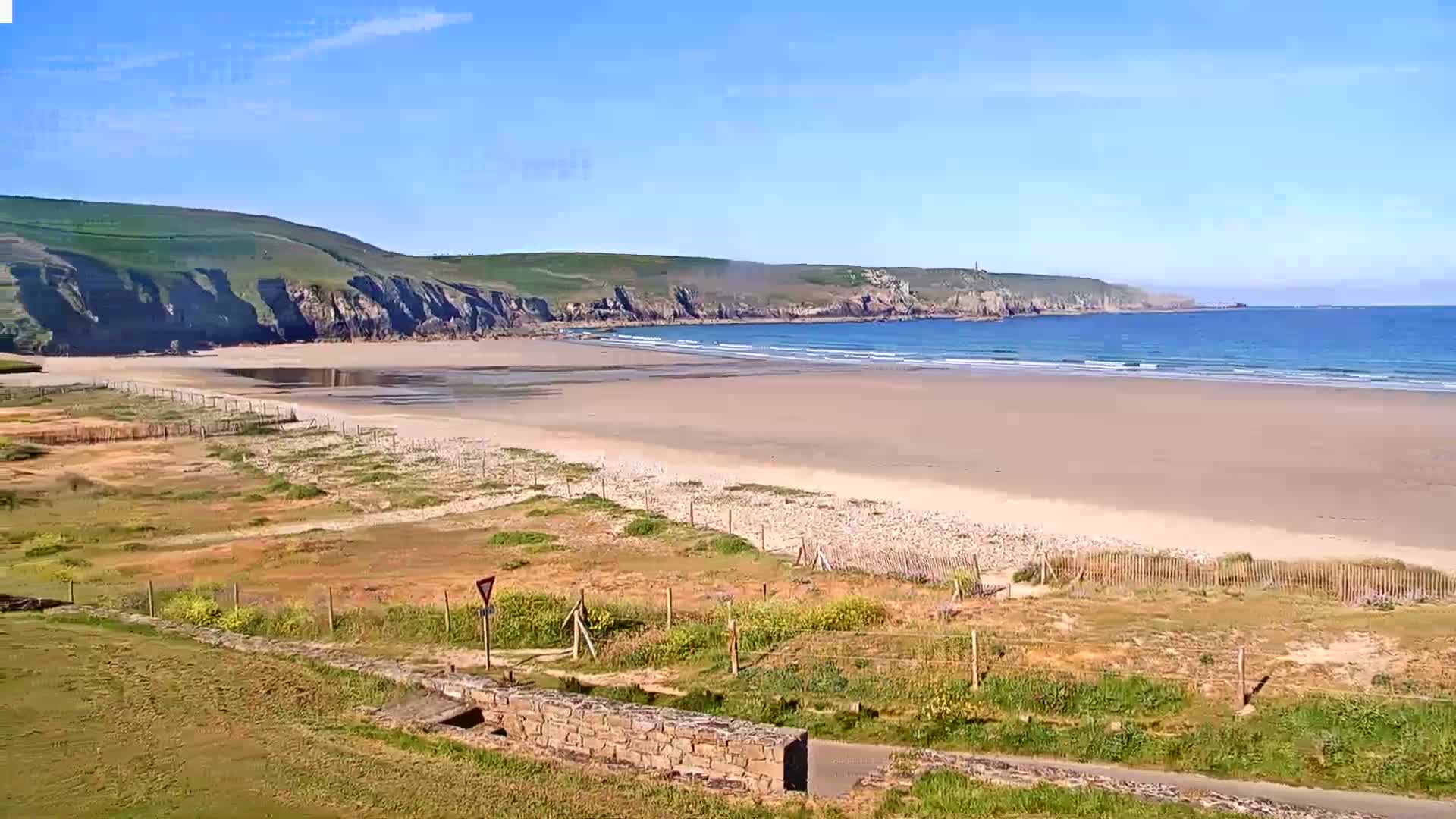 A sandy beach stretches out to a calm ocean under a clear blue sky, backed by green cliffs.