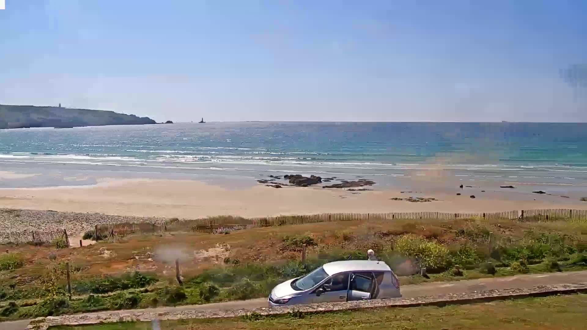 A silver car is parked near a sandy beach with light waves lapping the shore under a clear, sunny sky.