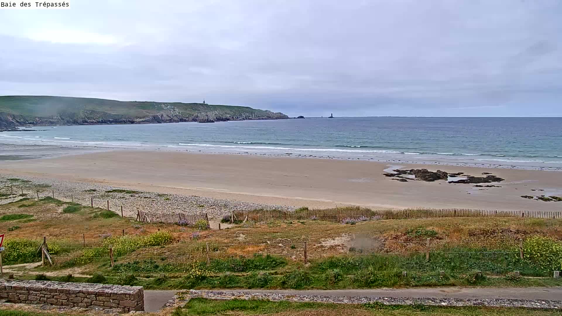 A sandy beach with gentle waves stretches toward a calm ocean under an overcast sky, backed by grassy dunes and a distant headland.