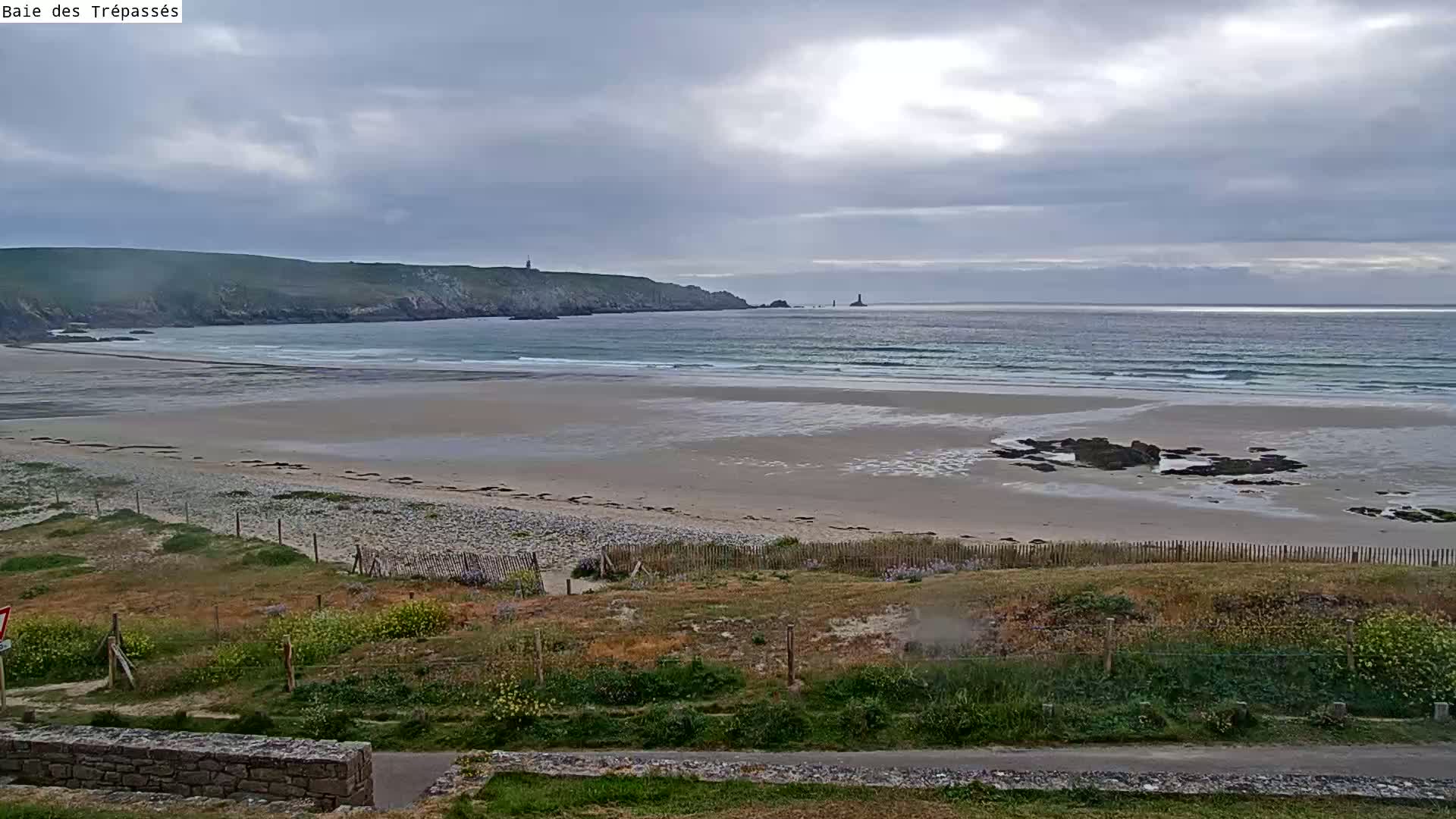 A long sandy beach with small waves under a cloudy sky is visible from a grassy overlook, featuring a rocky outcrop near the shore and a distant coastline with what appears to be a lighthouse.