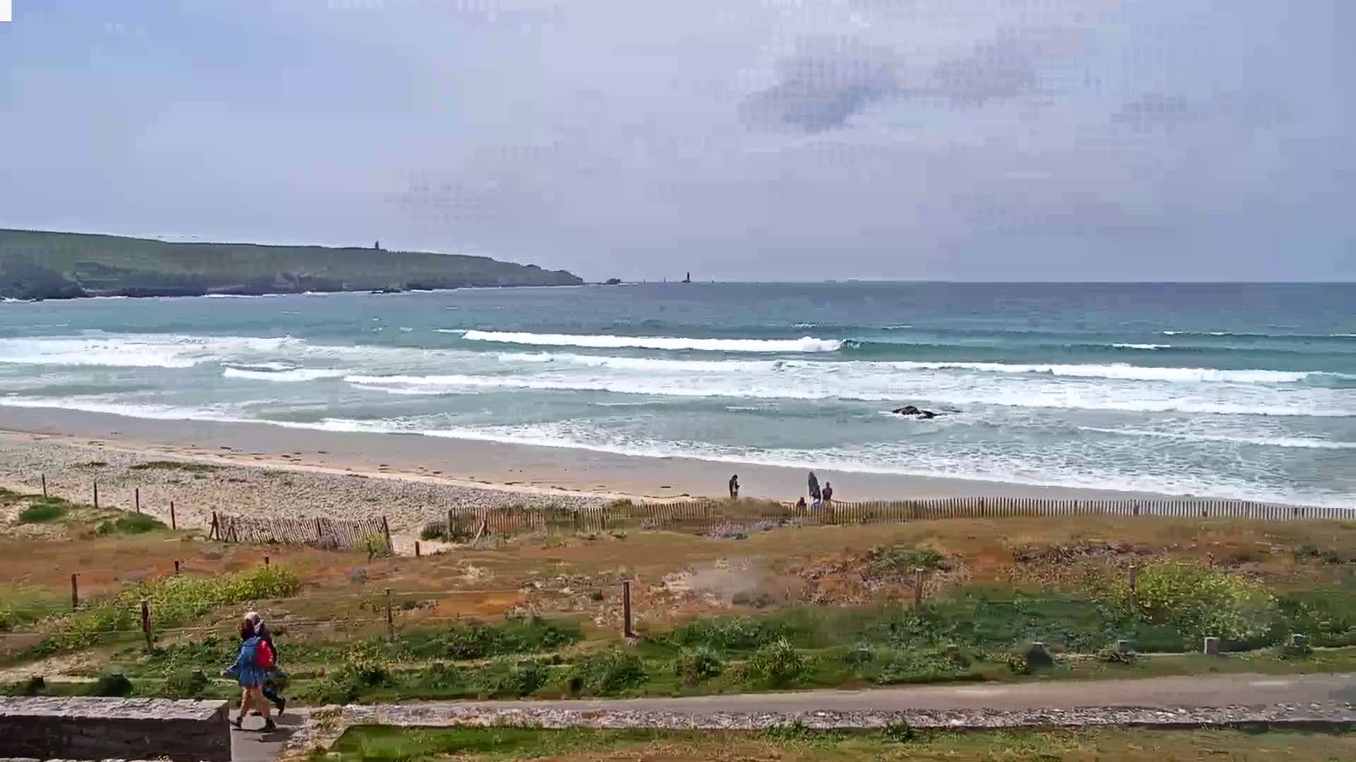 A sandy beach with gentle waves under a partly cloudy sky, a few people are visible on the beach and walking near the dunes.