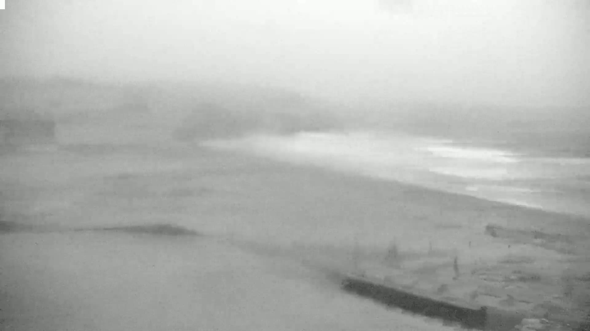 A foggy, overcast day reveals a beach with a partially visible ocean and indistinct structures in the foreground.