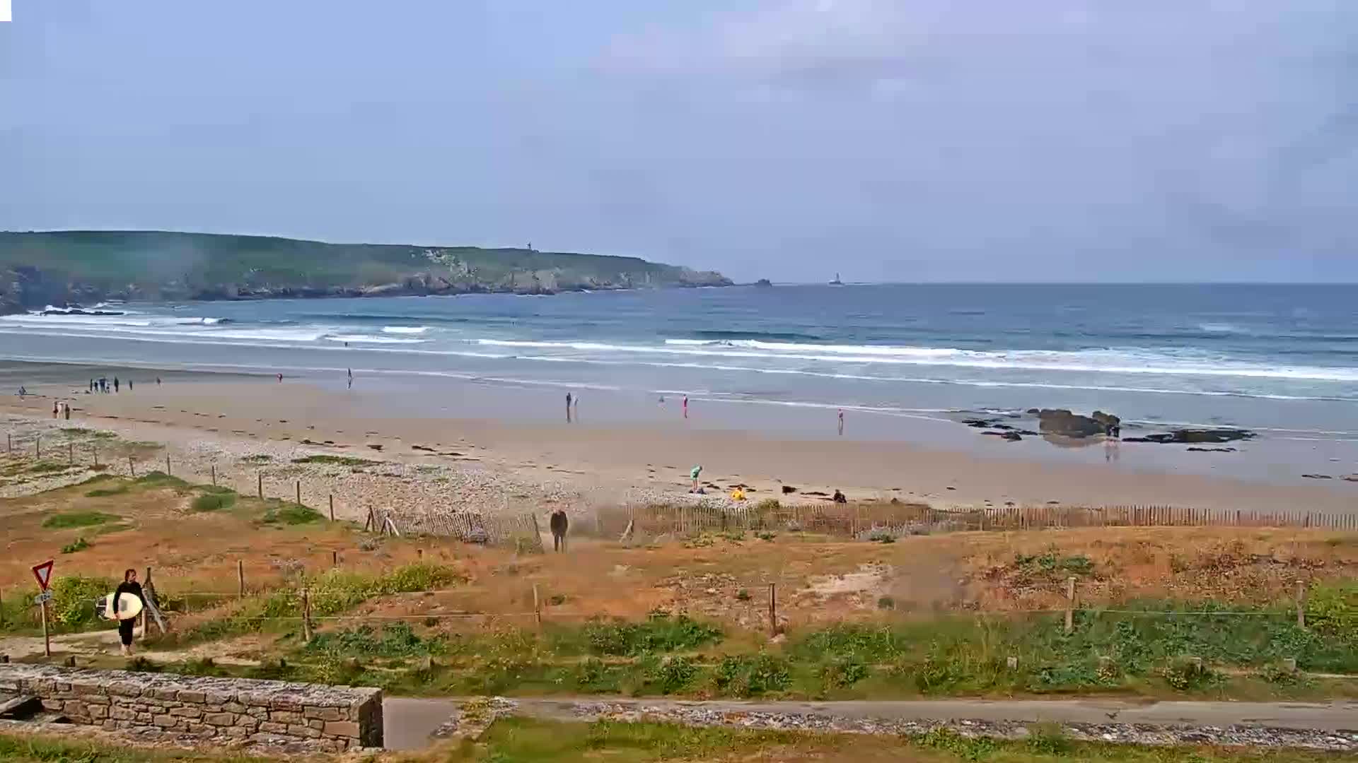 A sandy beach with small waves, a few people, and a rocky outcrop is visible, all under a mostly clear sky.