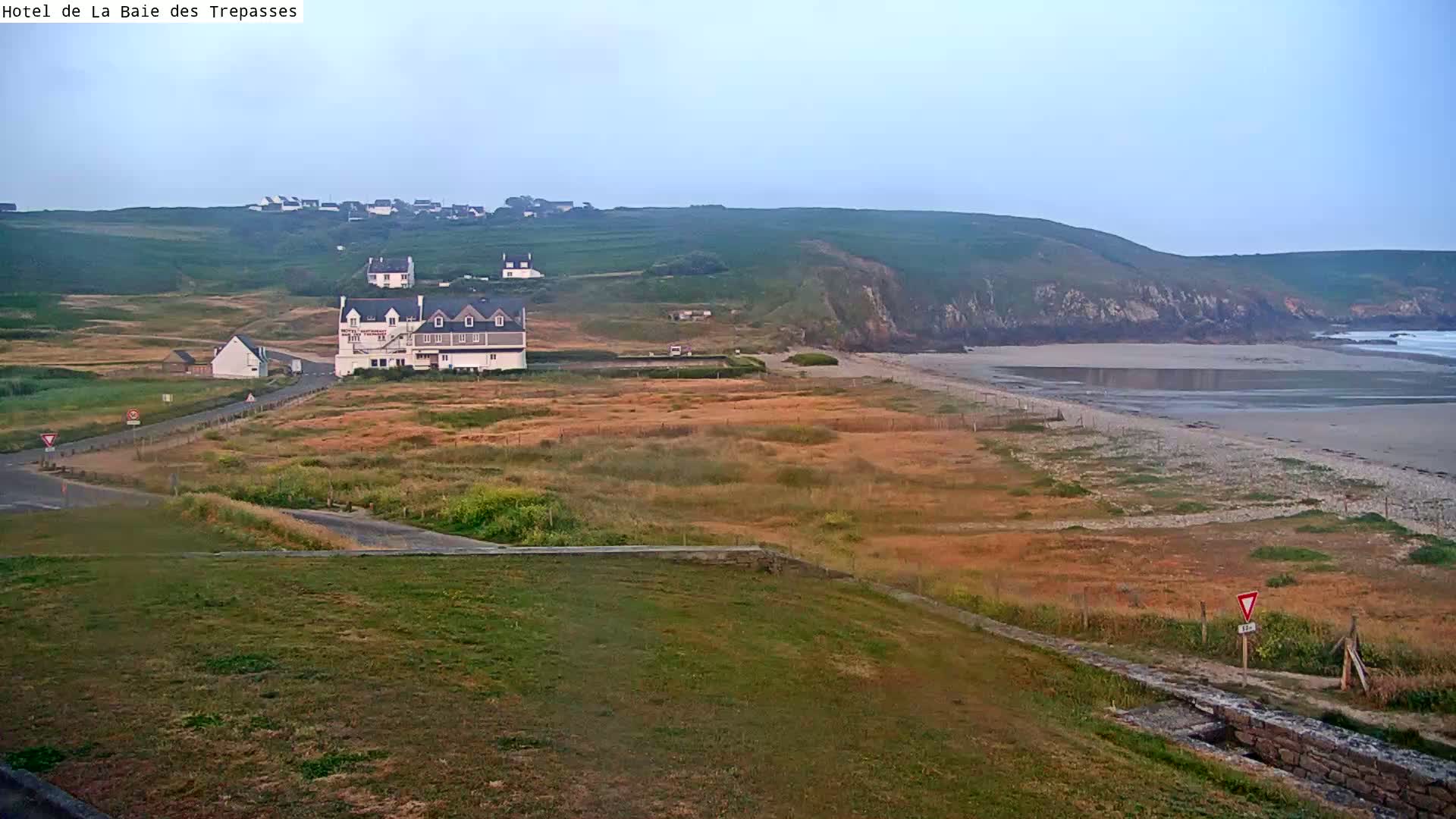 A coastal scene under a hazy sky shows a sandy beach, grassy areas, a large building, and a small village nestled on a hillside.