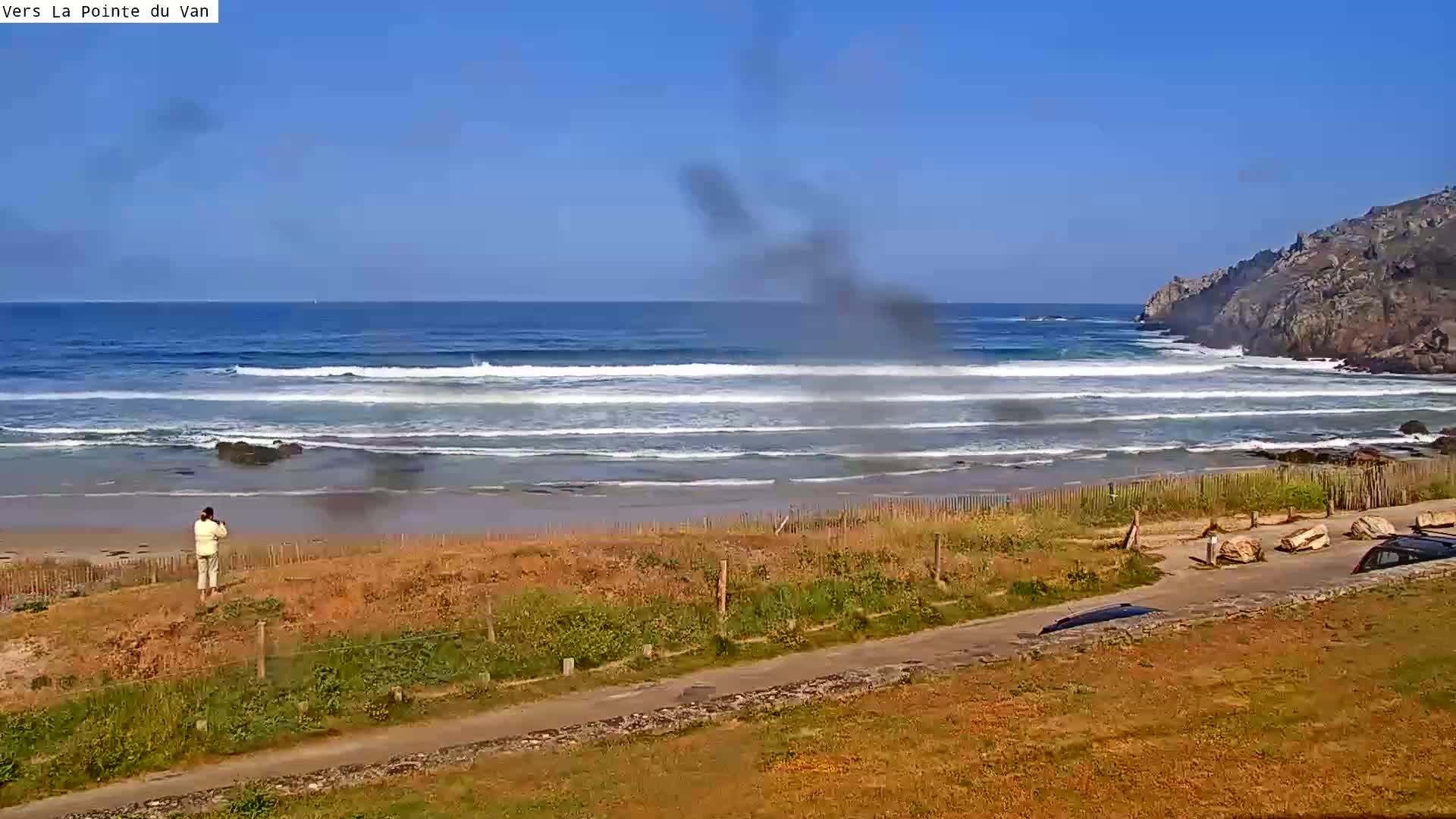 A sandy beach with small waves laps against the shore under a clear blue sky, with a person standing near the water's edge and cars parked nearby.