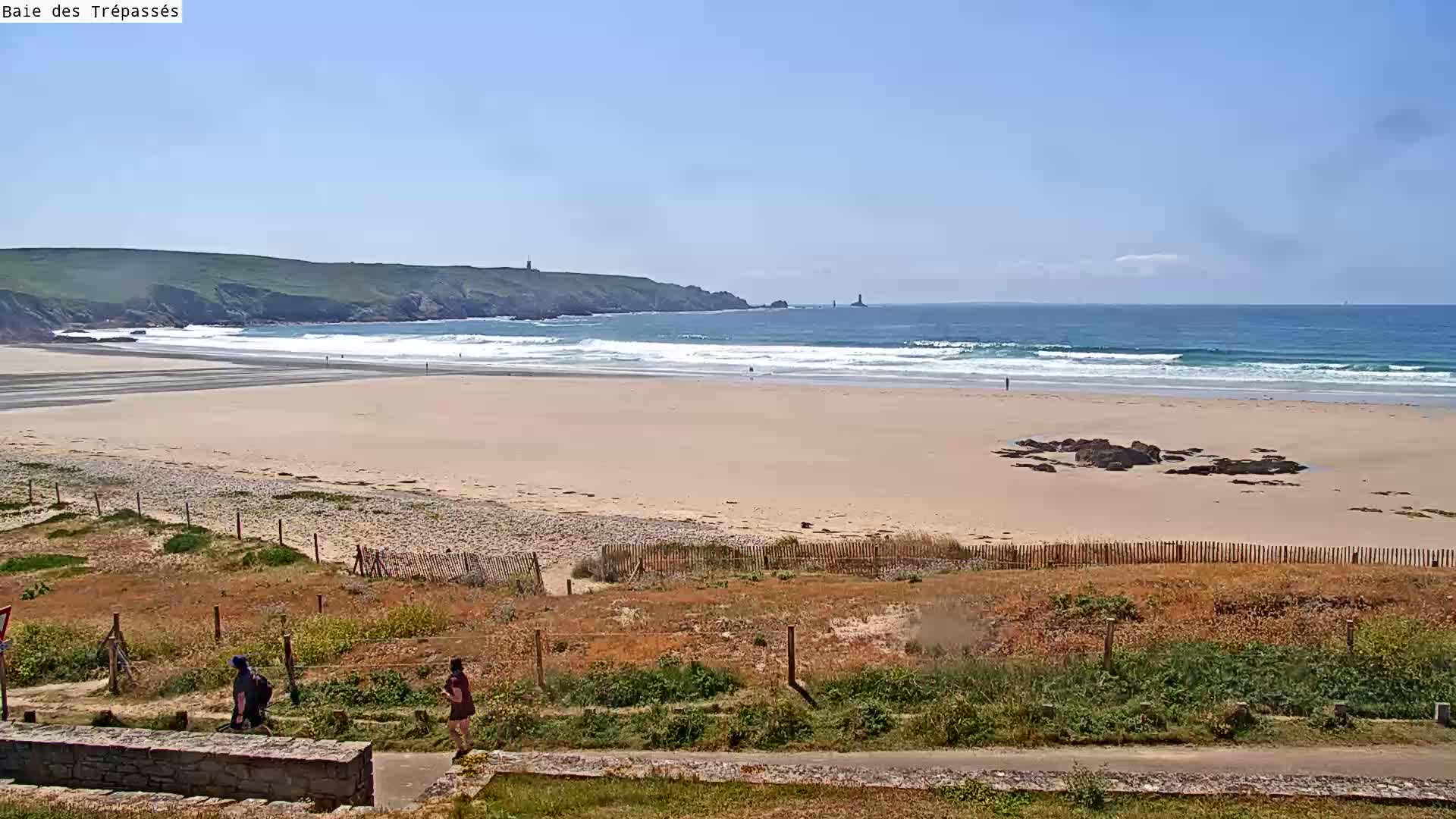 A sandy beach stretches to a blue ocean with small waves under a clear, sunny sky, and two people walk along the dunes in the foreground.