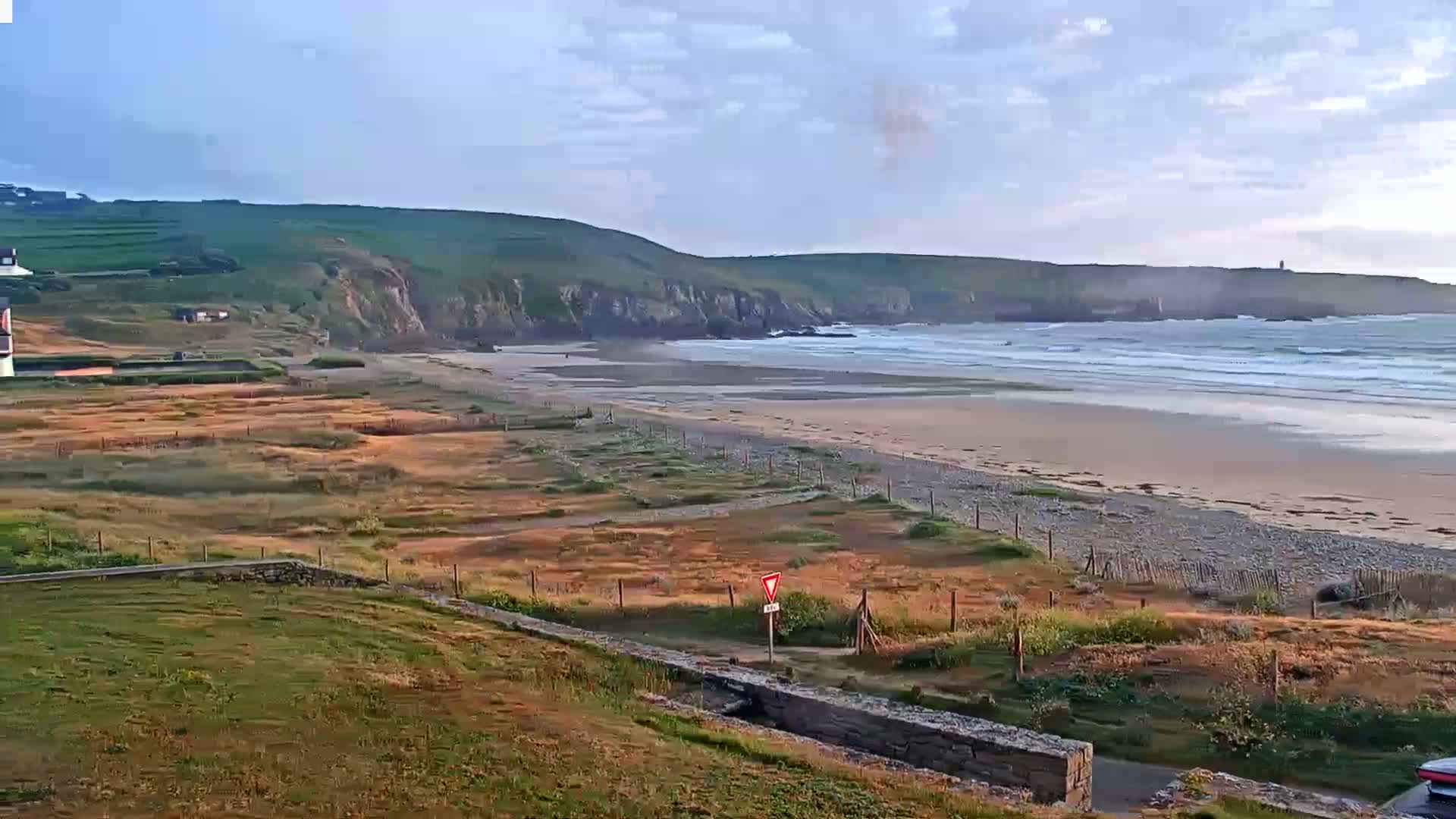 A sandy beach with small waves stretches along a rocky coastline under a partly cloudy sky.