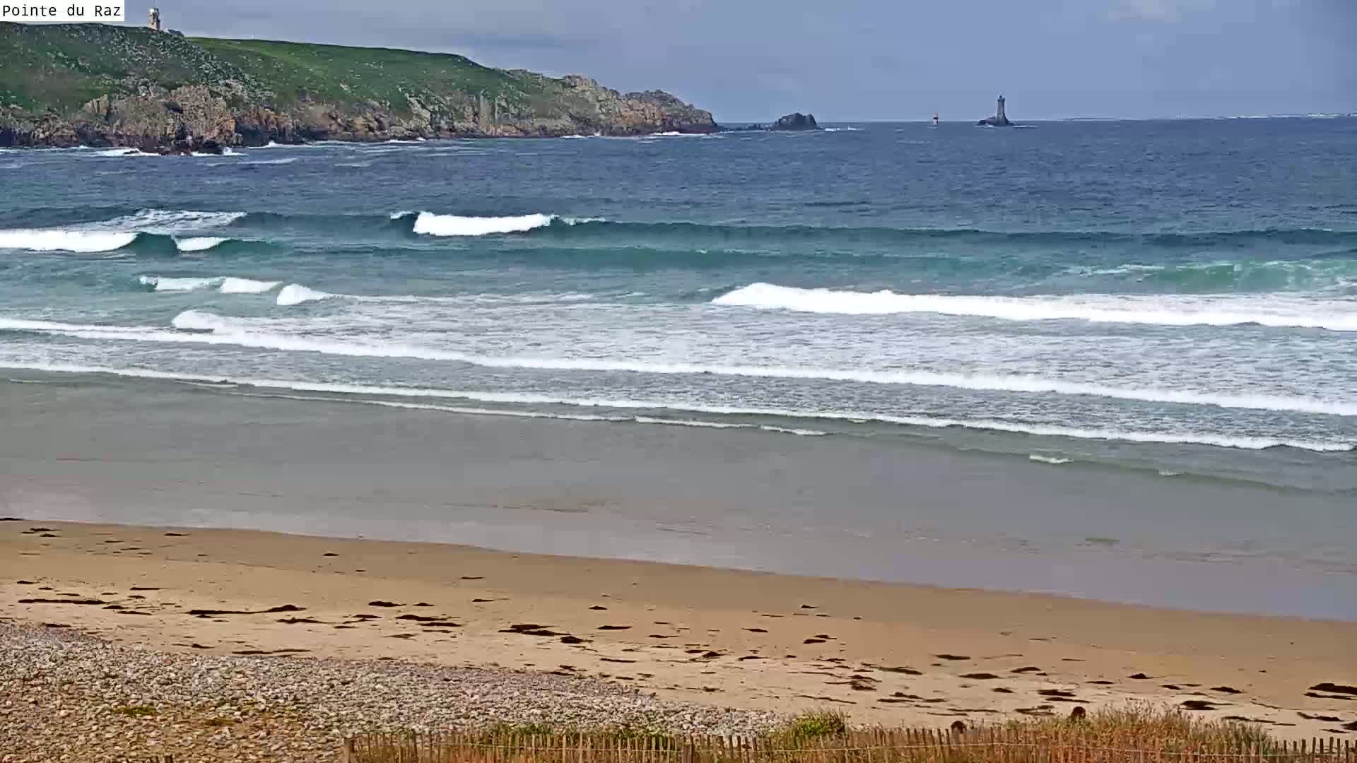 A sandy beach with small waves rolling onto the shore under an overcast sky, with a distant rocky coastline and a lighthouse visible on the horizon.