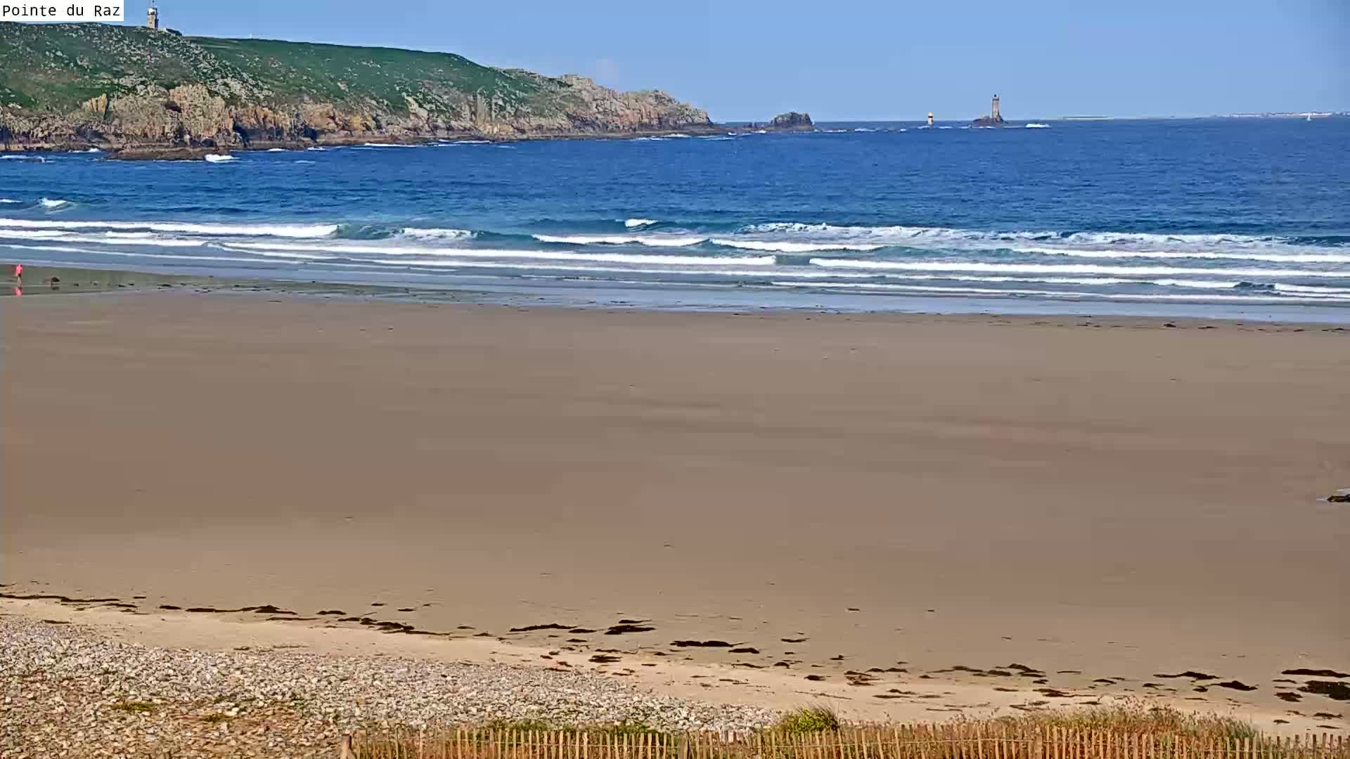 A wide sandy beach stretches to a calm ocean with small waves under a clear blue sky, and a distant lighthouse is visible on a rocky outcrop.