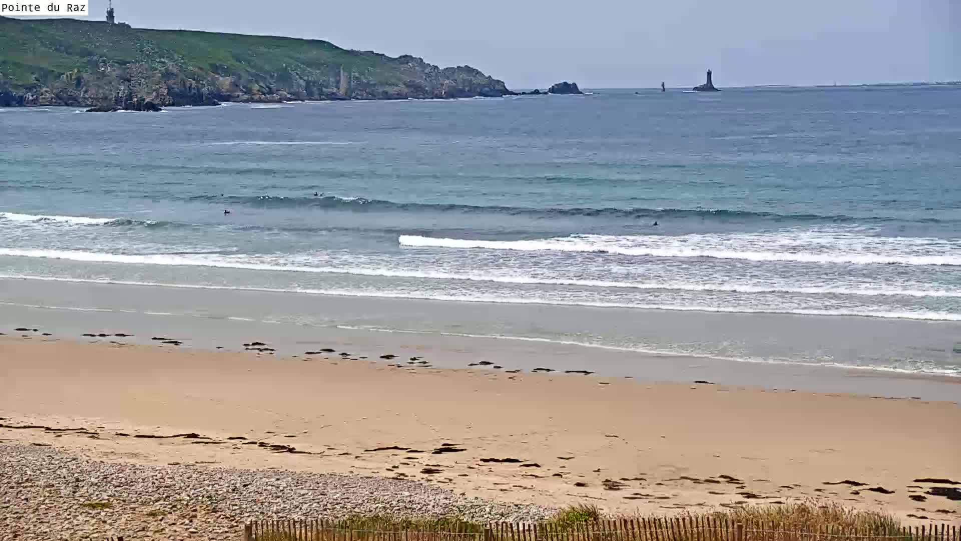 A sandy beach with small waves breaking gently, a few surfers in the water, and a distant rocky coastline under a mostly clear sky.