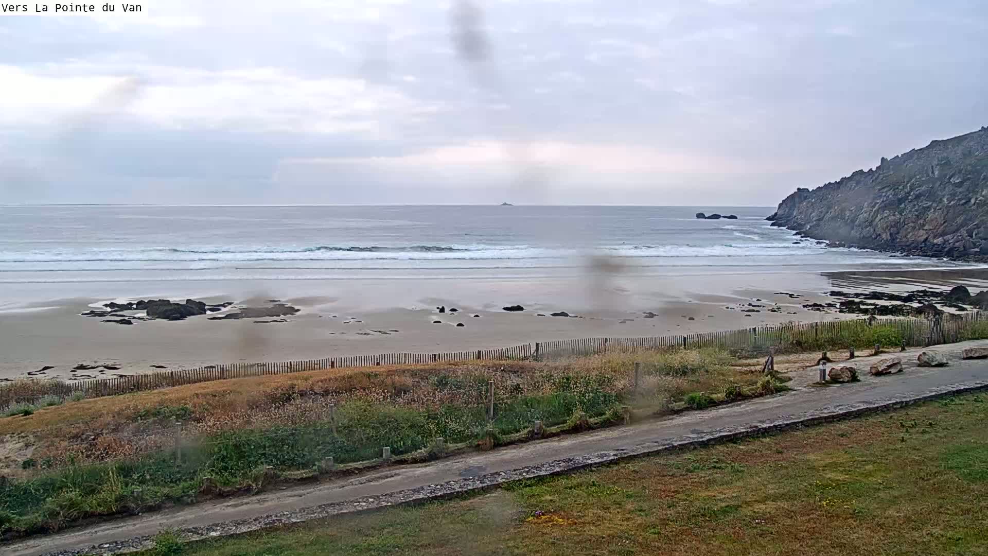 A sandy beach with small waves under an overcast sky is bordered by a rocky cliff and a grassy area with a road.
