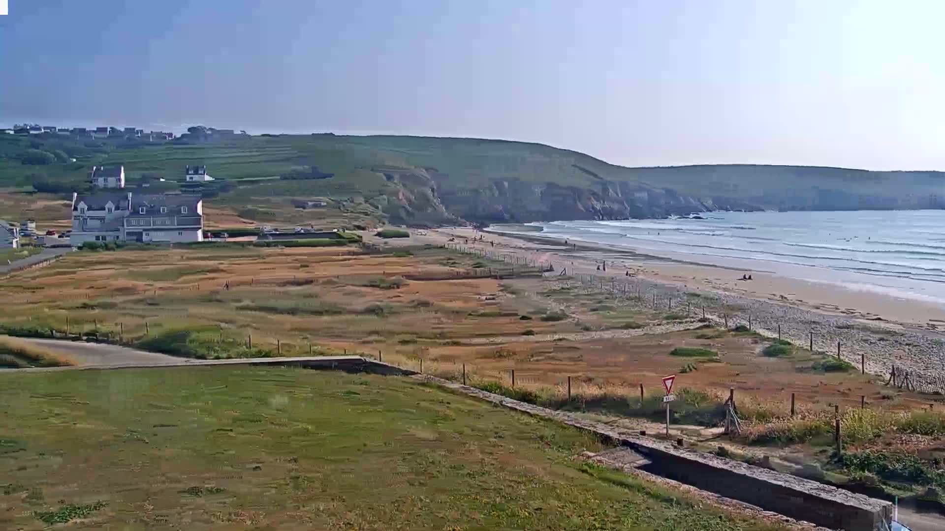 A sandy beach with gentle waves under a clear, sunny sky is situated below a grassy hill with a building and some sparse vegetation in the foreground.