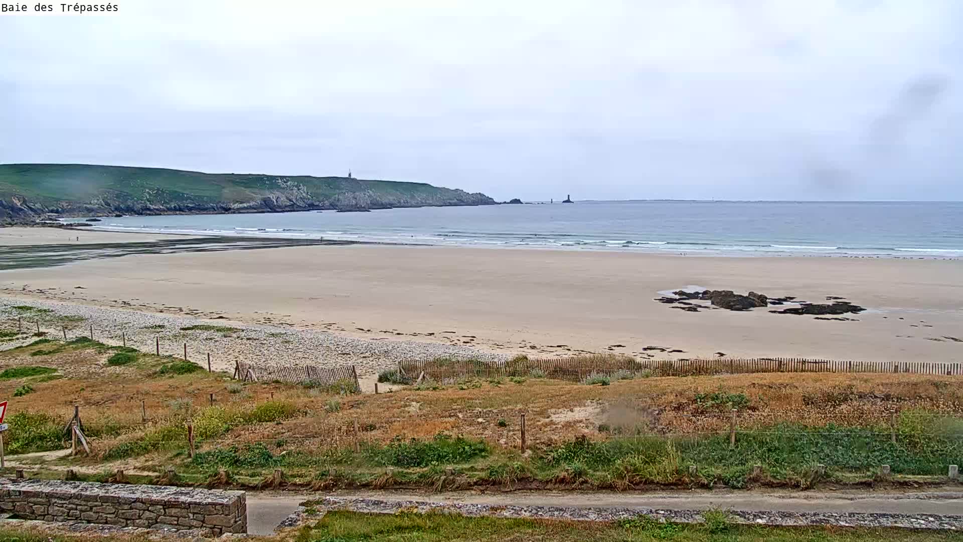 A long, sandy beach stretches towards a calm ocean under an overcast sky, backed by grassy hills and a distant headland.