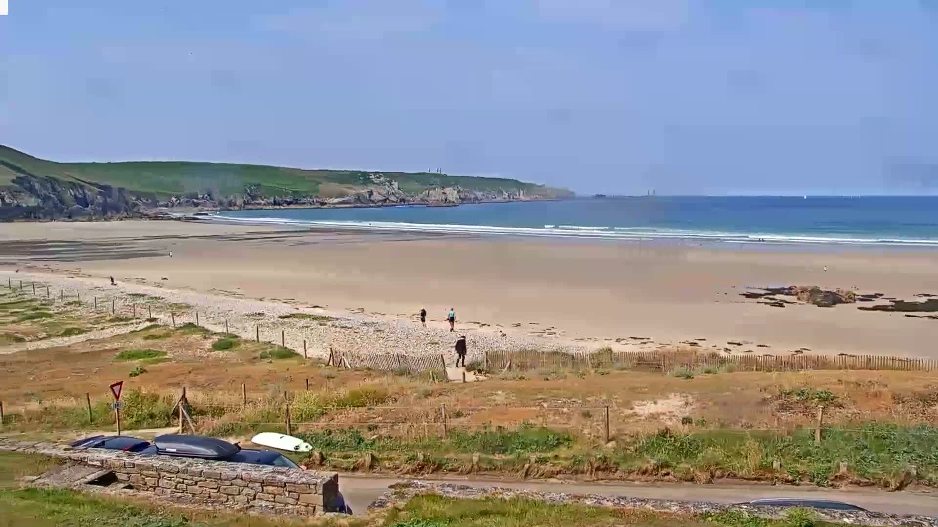 A sandy beach stretches along a calm ocean under a clear blue sky, with several people walking on the sand and cars parked nearby.