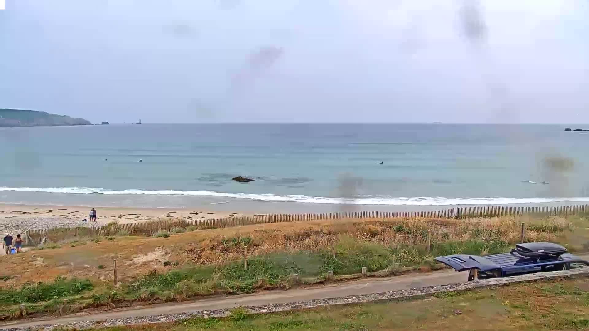 A sandy beach with a few people and surfers in the ocean under an overcast sky.