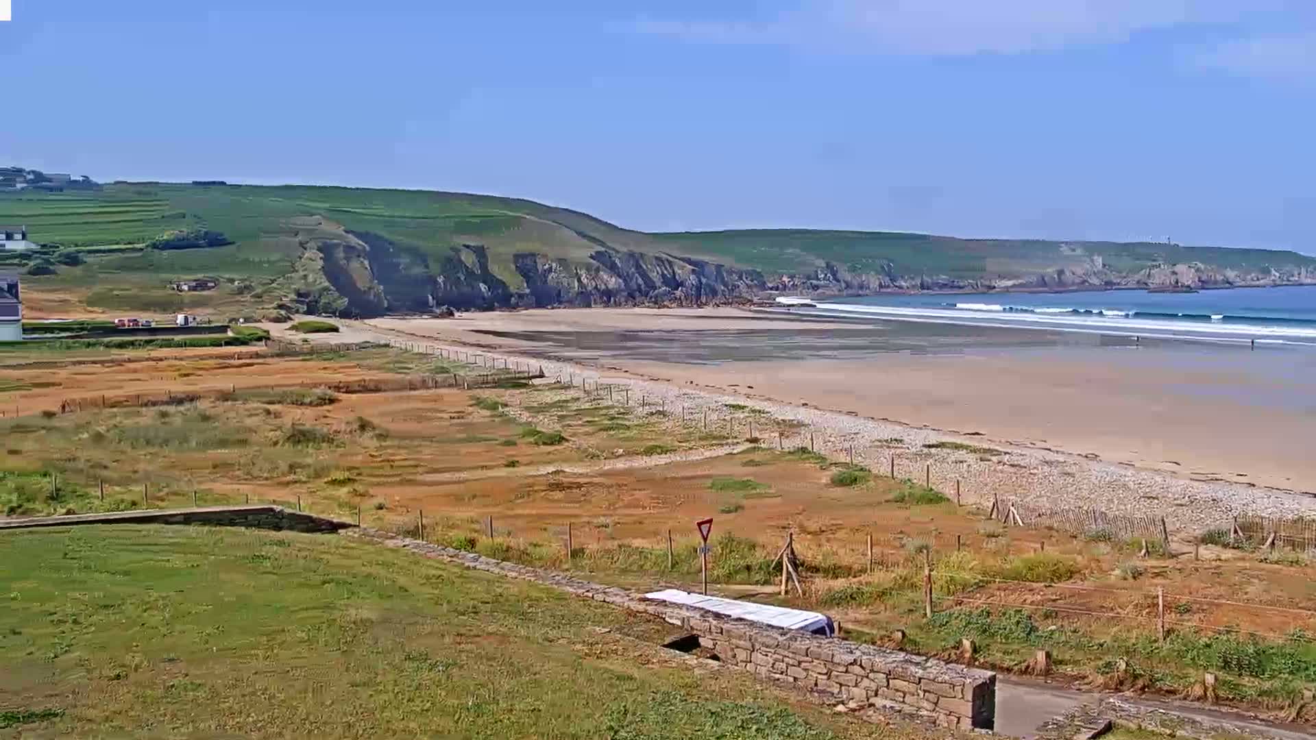 A sandy beach stretches along a rocky coastline under a clear blue sky, with small waves rolling in the ocean.