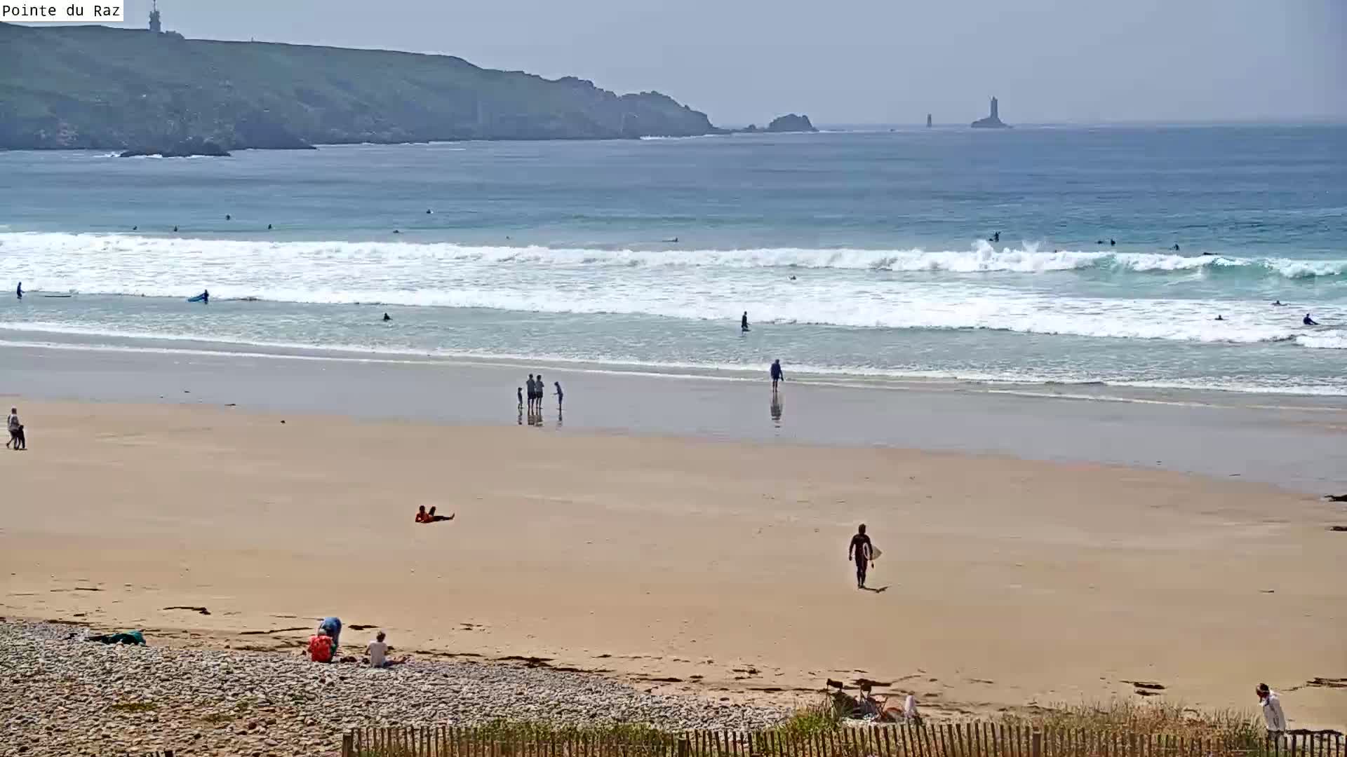 A sandy beach with several people sunbathing, walking, and surfing, under a mostly clear sky, features rolling waves and a distant rocky coastline with a lighthouse.