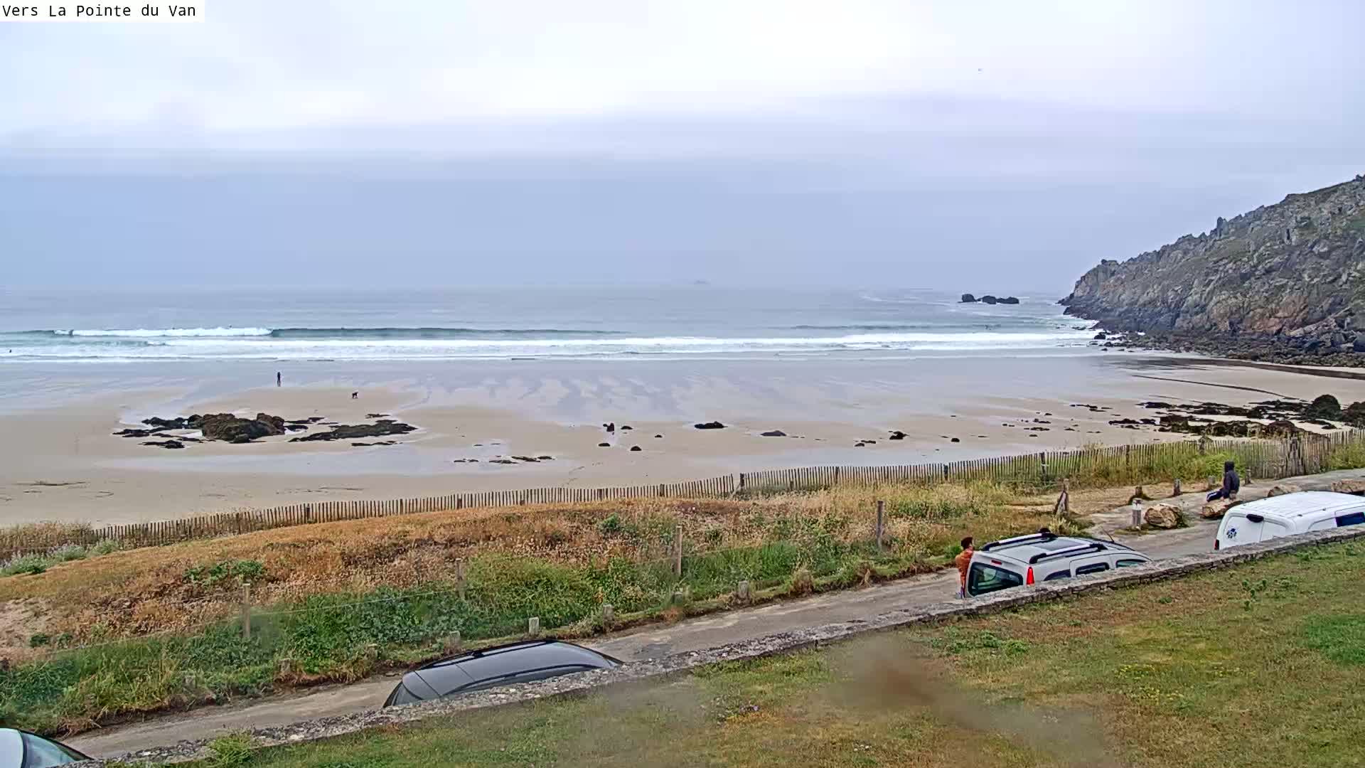 A sandy beach with small waves under an overcast sky is seen from a roadside vantage point, with several vehicles parked nearby and people standing near them.