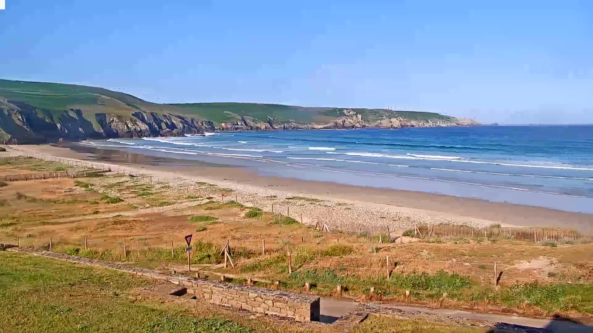 A sandy beach with gentle waves laps a shoreline bordered by grassy hills under a clear blue sky.