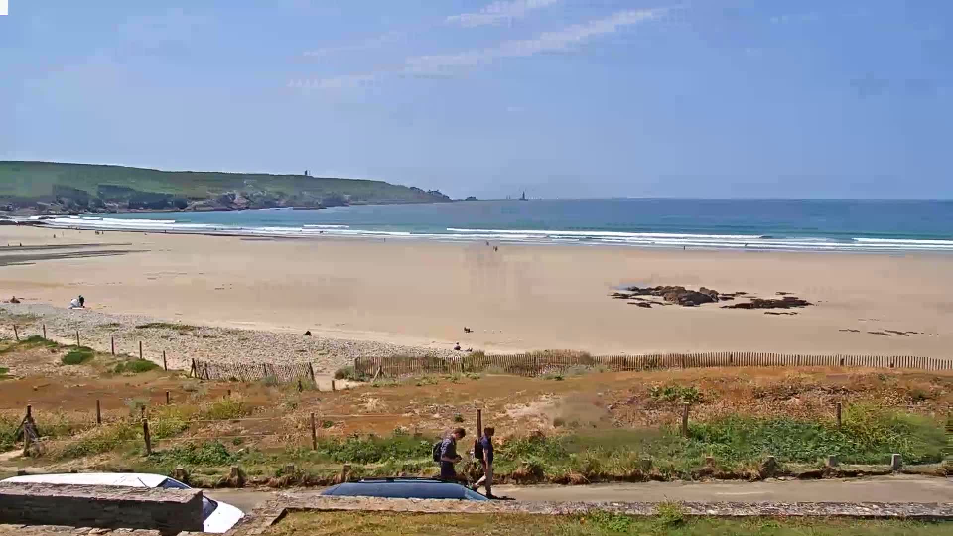 Two people walk along a road overlooking a sandy beach with gentle waves under a clear blue sky.