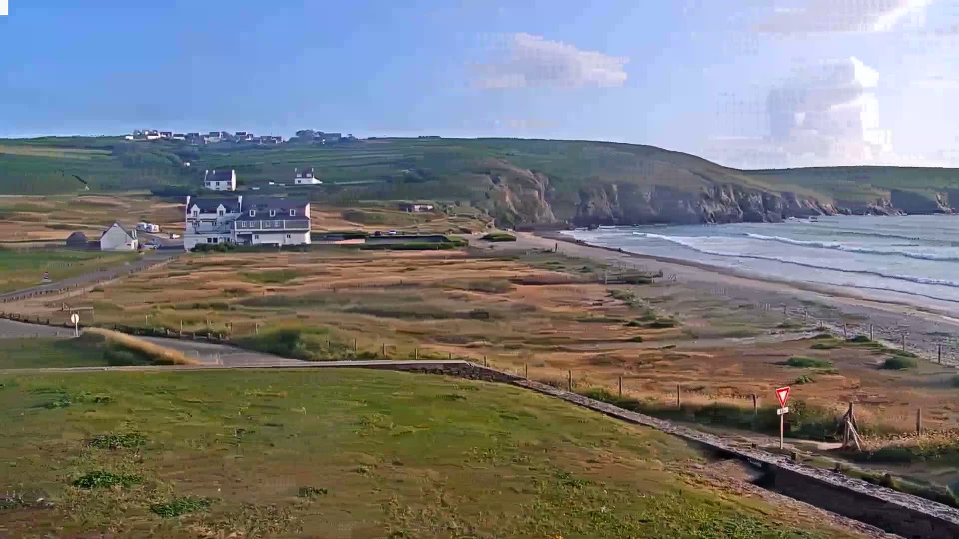 A coastal scene under a mostly clear sky shows a sandy beach with gentle waves, a small village nestled on a hillside, and a large building near the beach.