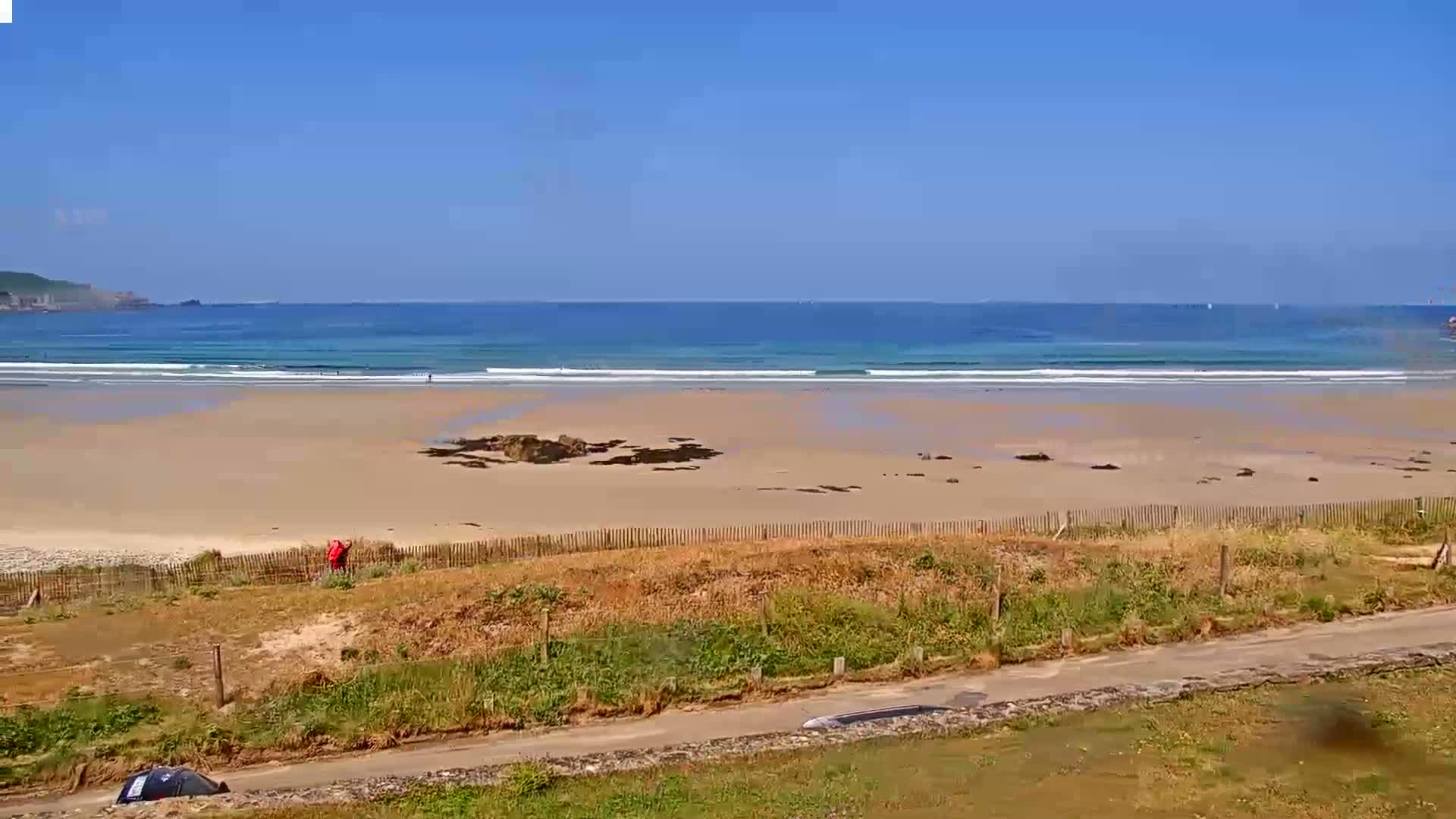A sandy beach with gentle waves under a clear blue sky is viewed from a slightly elevated vantage point, showing a person on the beach and a road in the foreground.