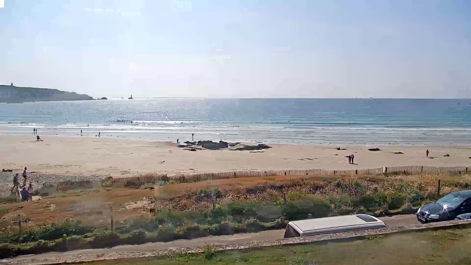 A sandy beach with a few people on it stretches along a calm ocean under a sunny sky, viewed from an elevated position above a grassy area with parked vehicles.