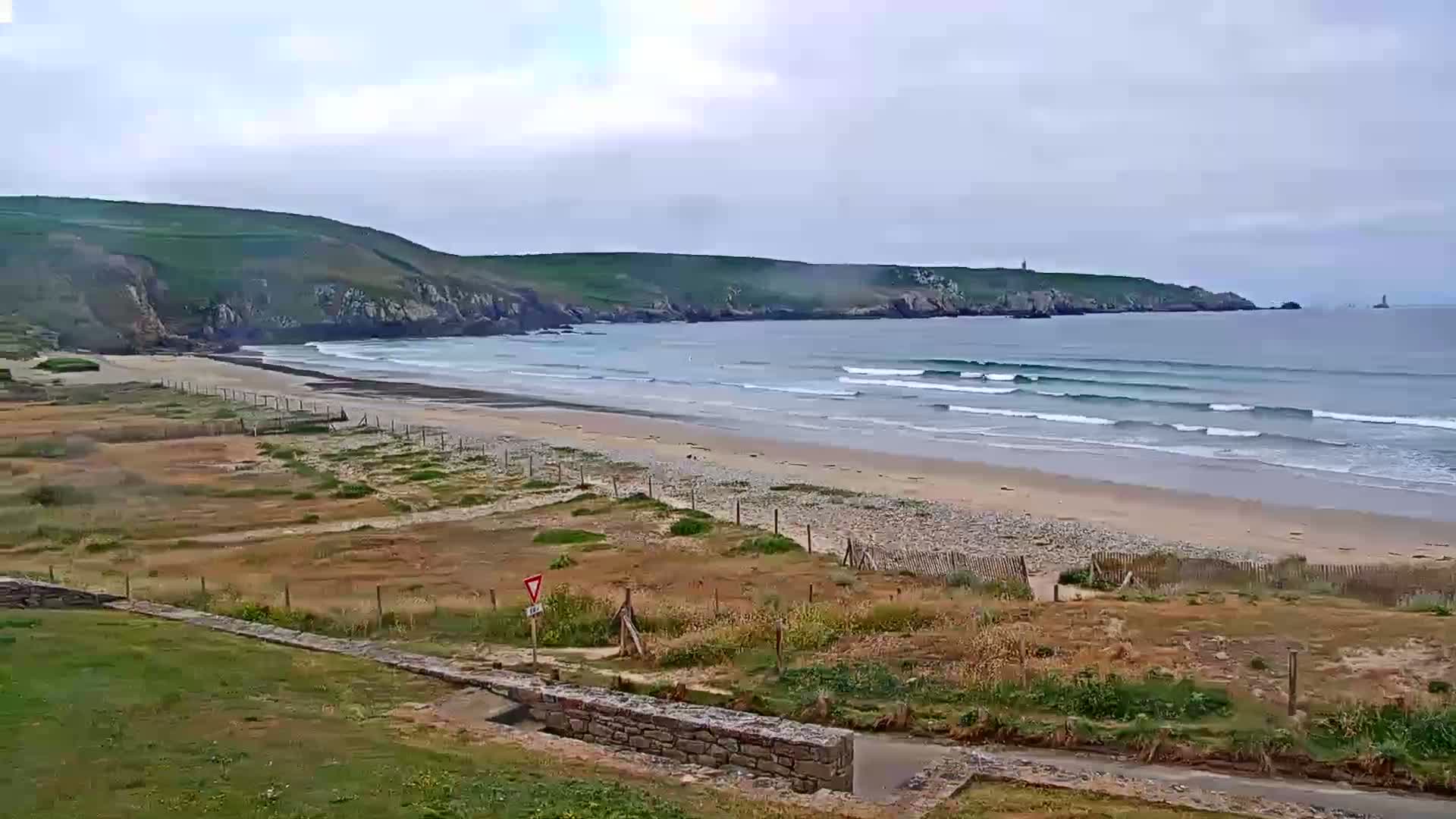 A sandy beach with gentle waves rolls towards a rocky, green headland under an overcast sky.